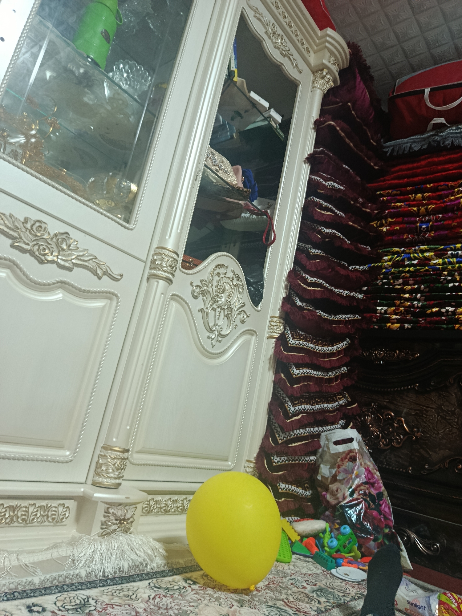 In a room with textured ceilings, a cream-colored wardrobe with ornate gold trim stands prominently. The wardrobe has glass doors revealing shelves stocked with items, including a green thermos and decorative glassware. To the right of the wardrobe, a stack of folded maroon and red prayer mats with intricate patterns and fringed edges is visible, topped by a red bag. In the foreground, a bright yellow balloon rests on a patterned rug, near a collection of colorful children's toys and a plastic bag with a floral print. A white tassel hangs from the wardrobe base.