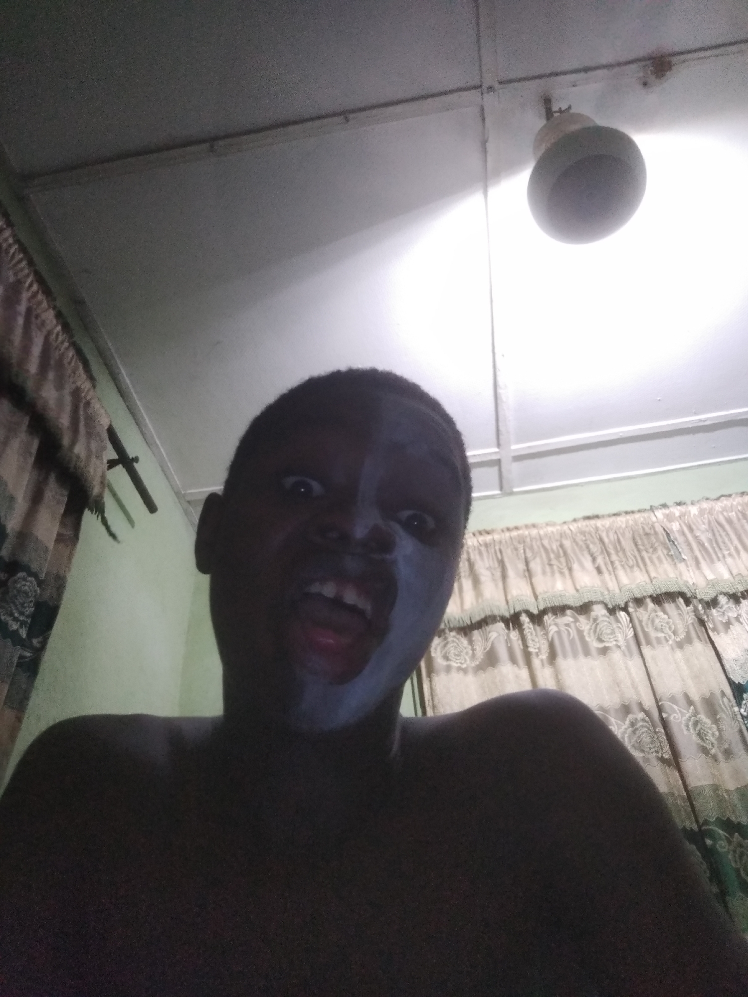 A low-angle close-up shot of a person with dark skin and face paint. The person is looking up and opening their mouth. The background includes a ceiling, a light fixture, curtains, and a wall. The location is in a room, possibly in a house or apartment in Shangisha, Nigeria.