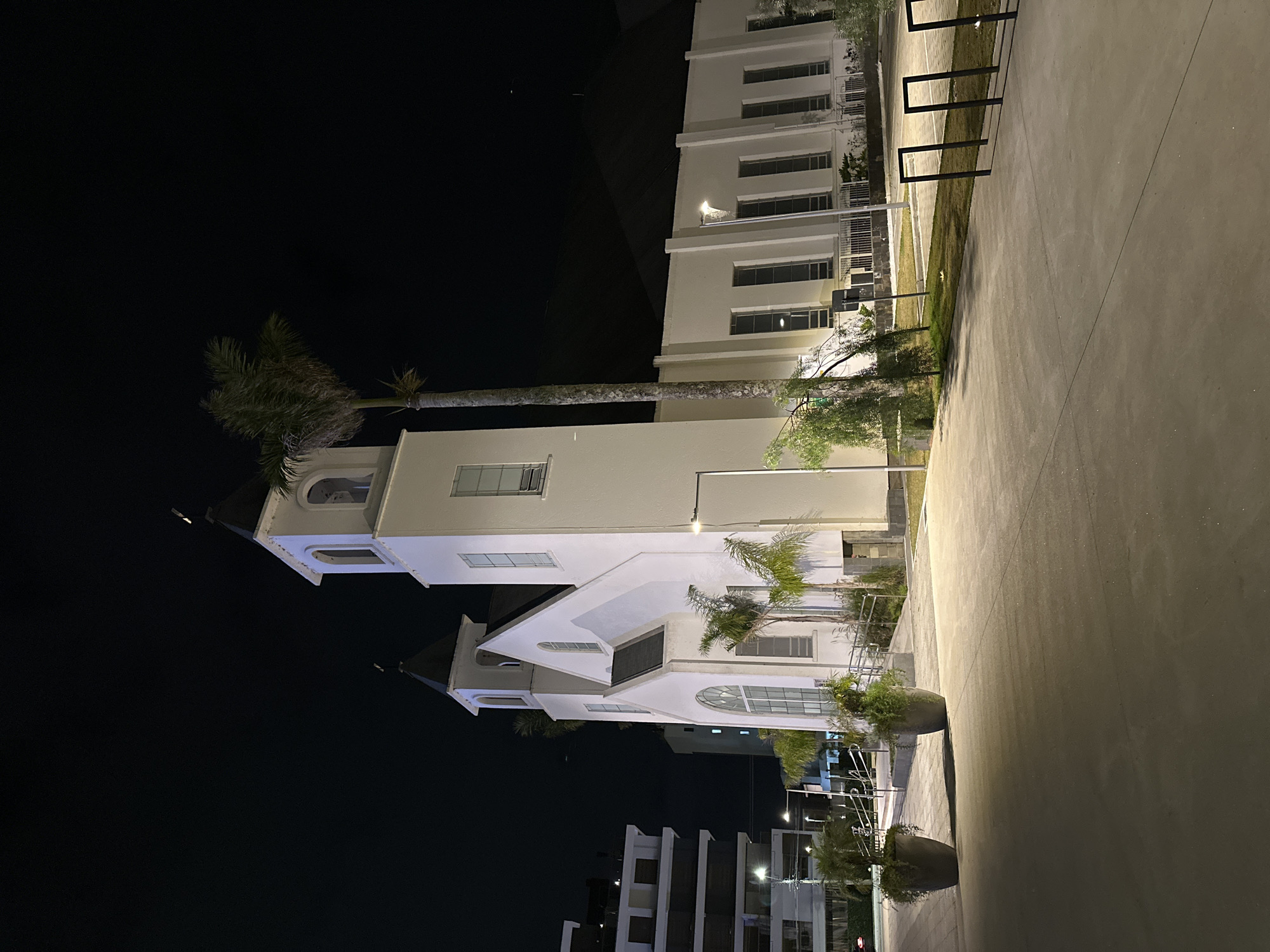 The image shows a white church with a dark roof and two steeples at night in the city of Florianópolis, Brazil. The church is illuminated by artificial lights, casting shadows on the surrounding paved area. In front of the church, there are two large planters with greenery, and several palm trees. To the left of the church, there is a modern, multi-story building. The sky is dark and clear. The scene appears to be a public space, possibly a plaza or promenade, with the church as the central architectural feature.