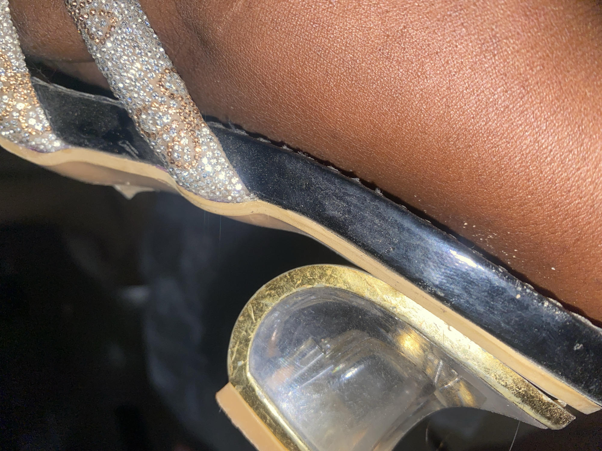 The image is a close-up shot of a person's foot wearing a jeweled sandal. The sandal has a clear heel with gold trim, a black sole, and a strap adorned with rhinestones in silver and rose gold. The person's skin tone is dark brown. The background is dark and out of focus, making it impossible to determine the location within Jauro Yinu, Nigeria. No actions or interactions are depicted.