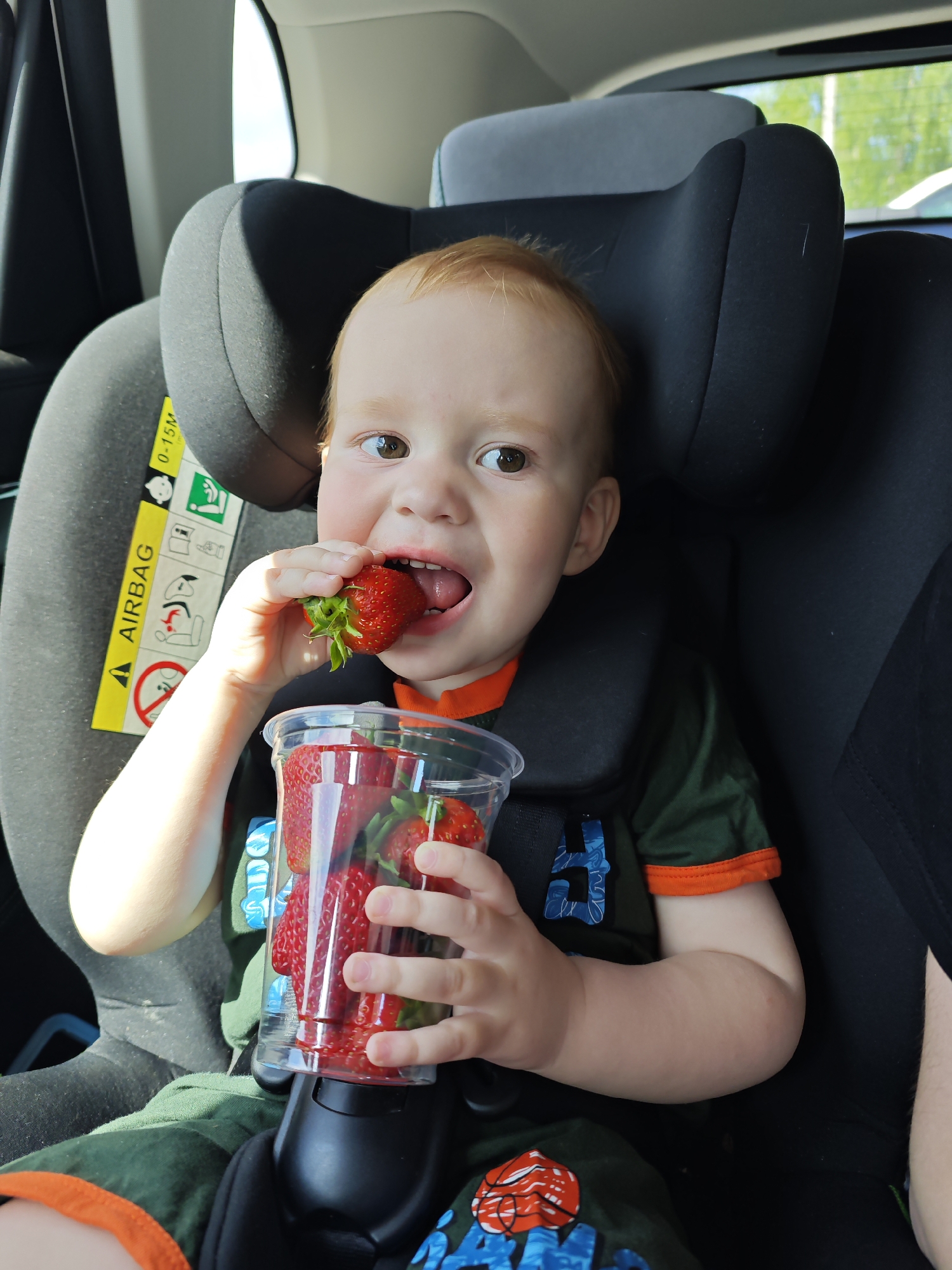 A toddler with red hair is sitting in a grey car seat, secured by a black harness, holding a clear plastic cup full of strawberries. He is eating one of the strawberries, his tongue slightly visible. He is wearing a dark green t-shirt with orange trim and a blue and orange design. The car seat has a yellow "AIRBAG" warning sticker. The location is Chaikovsky, Russia.