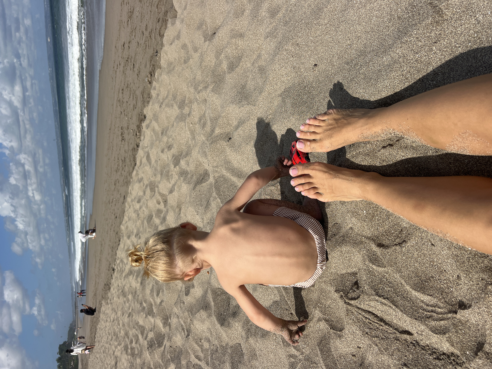 The image depicts a bright, sunny day at a sandy beach, likely in North Kuta, Indonesia. The scene is dominated by the textured sand, with waves gently rolling in from the ocean on the left side of the frame. The water appears calm with a white surf line.
In the foreground, a young child with blonde hair, tied in a bun, is playing in the sand. The child is facing away from the camera, engrossed in their activity. They are wearing striped shorts. A small red toy car is visible near the child's hands, suggesting they are playing with it.
In the upper right quadrant of the image, a person's legs and feet are visible, suggesting they are sitting or lying in the sand close to the child. The person has painted toenails with a light pink color. Their legs are also covered in sand. The sunlight casts shadows on the sand, indicating the time of day is likely late morning or afternoon.
In the background, further down the beach, several other people can be seen, some walking and some seemingly sitting or playing. They are distant figures, adding to the sense of a relaxed beach atmosphere. The sky is partly cloudy with some white clouds visible.
There is no visible text in the image. The overall mood is one of a peaceful and enjoyable day at the beach.