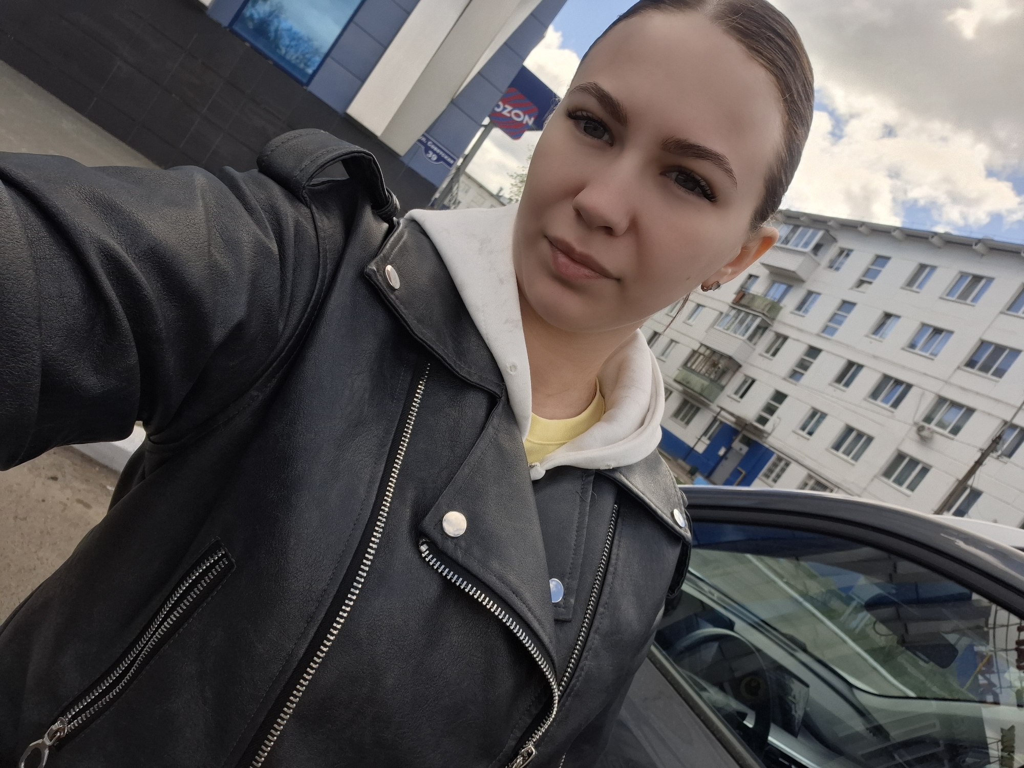 A woman in a black leather jacket over a white hoodie and yellow shirt takes a selfie outdoors. Behind her, partially visible, is a gray car. In the background is a multi-story white building with repeating window patterns and a sign displaying "OZON." The location is Chaikovsky, Russia.