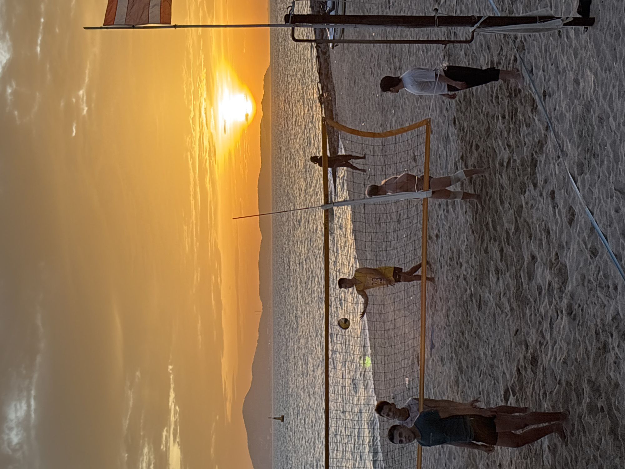 An outdoor, eye-level, full shot captures a beach volleyball game at sunset in Pattaya City, Thailand. The sun, a bright orb low on the horizon, casts a warm, orange glow across the sky and illuminates the choppy sea. A mountain range is visible in the distance, silhouetted against the colorful sky. Several men are playing volleyball on a sand court. Two players are on one side of the net, and three players are on the other. One player in a yellow jersey with the number 23 is mid-air, hitting the ball. The other players are positioned around the court, some watching the ball, others preparing to play. A volleyball net stretches across the court, dividing the players. In the foreground, a volleyball pole with a flag and some attached ropes stands to the right of the frame. The sand is unevenly textured with visible footprints and debris.