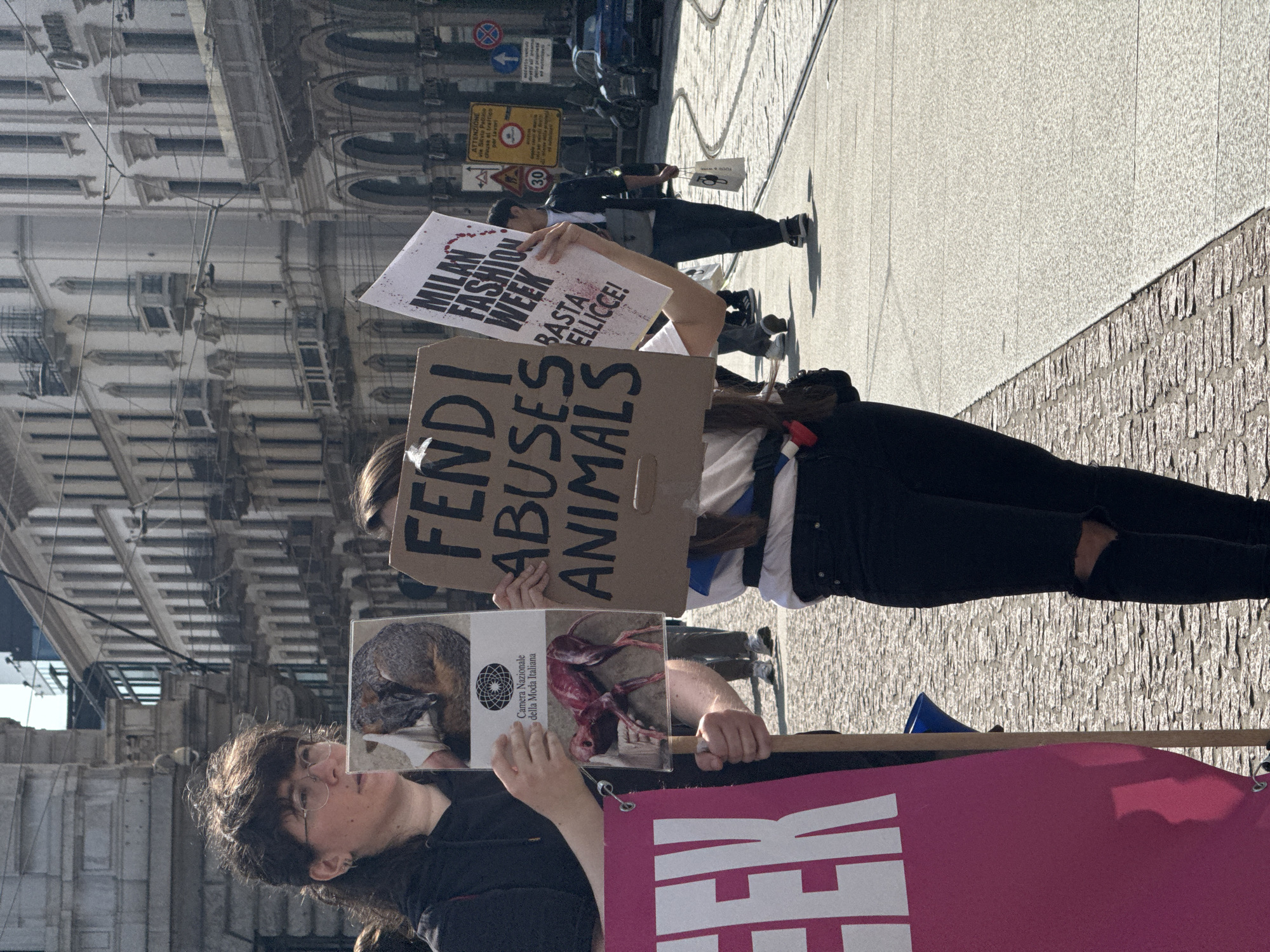 The image captures an animal rights protest taking place in Milan, Italy, during Milan Fashion Week. The protesters are holding signs and banners with messages advocating against animal cruelty. Some signs read "FENDI ABUSES ANIMALS" and "MILAN FASHION WEEK BASTA PELLICCE!", suggesting their focus is on luxury brands using animal fur. One protester holds an image of a dead animal. In the background, the architectural details of the city of Milan are visible, including signage with a speed limit. A portion of a traffic sign warning of roadwork is also visible.