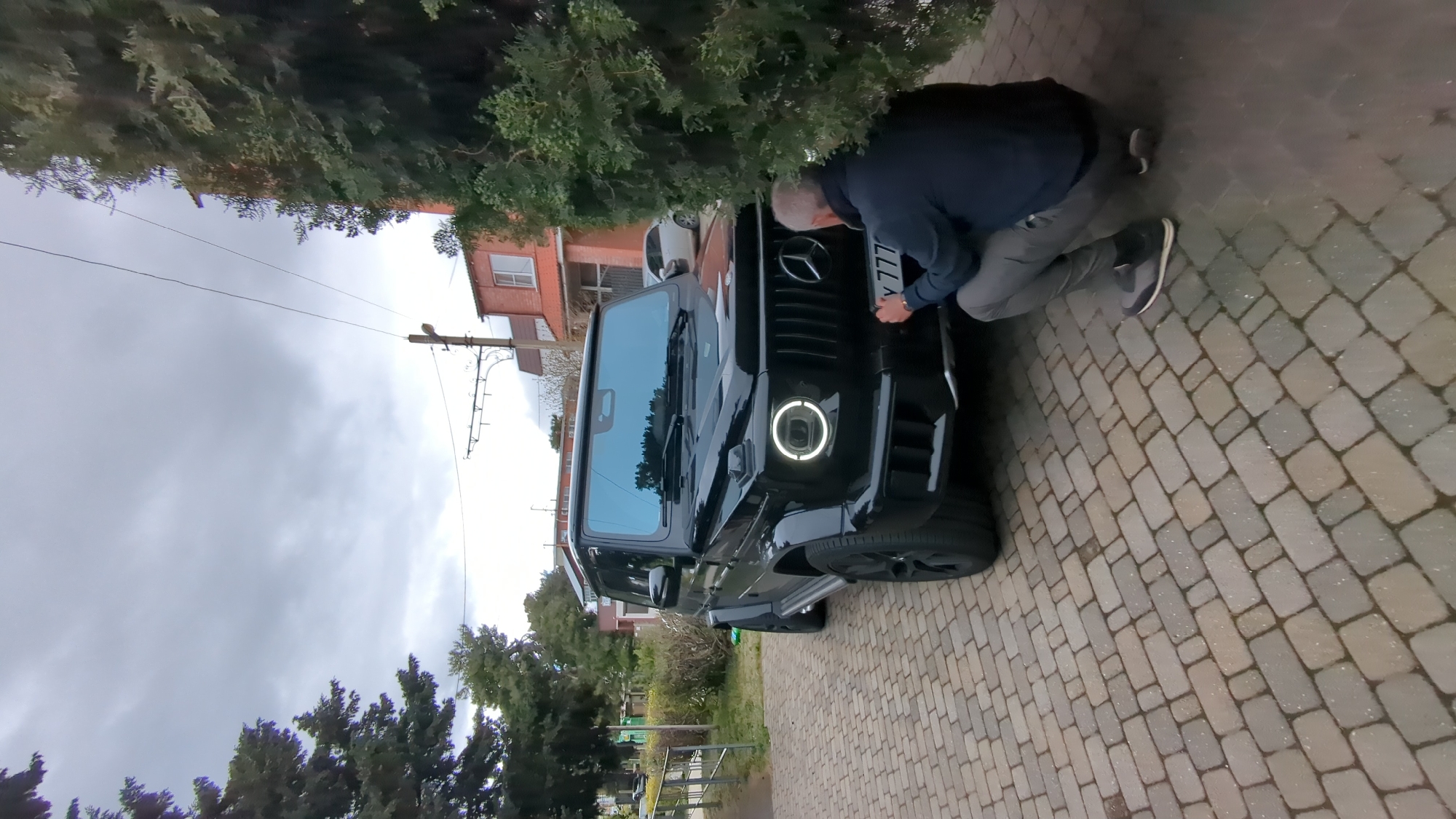 A person is attaching a license plate to a black Mercedes-Benz G-Class SUV. The license plate reads "Y 777". The vehicle is parked on a paved driveway with a brick pattern. Behind the vehicle is a residential building with red brick and white window frames. The scene appears to be outdoors on a cloudy day, likely in Gelendzhik, Russia. The presence of the license plate suggests the activity is focused on vehicle registration.