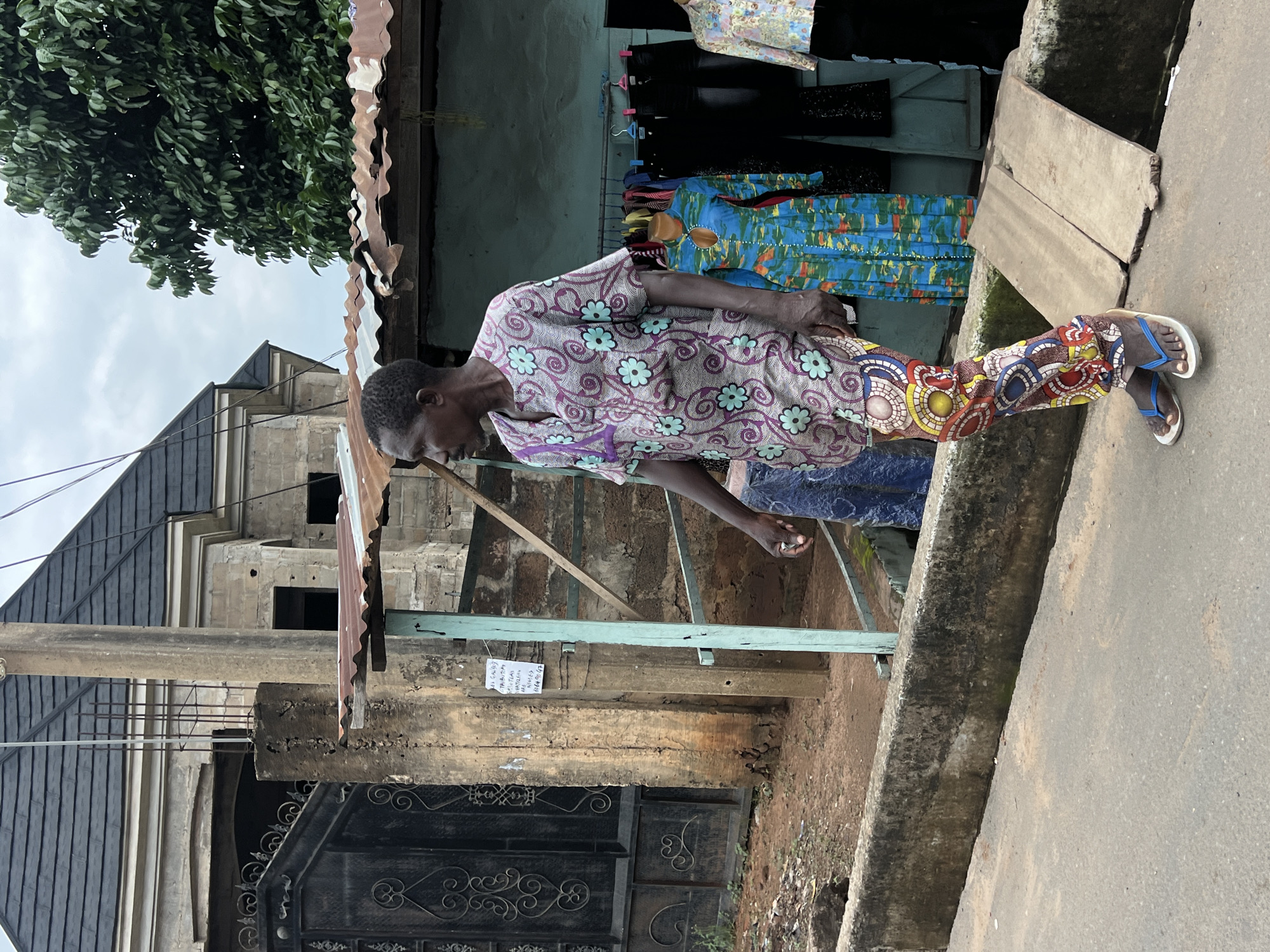 In Nkpor, Nigeria, a man is walking on the sidewalk in front of a small shop. The man is wearing a colorful, patterned outfit consisting of a tunic and pants, along with blue and white flip-flops. Behind him, the shop displays several articles of clothing, including a blue and green patterned dress on a mannequin and several pairs of black pants hanging on a rack. The shop has a corrugated metal roof, and a blue wooden frame supports an awning. To the left of the shop, a large, ornate black metal gate is visible, and beyond that, a building with a dark tiled roof and exposed rebar suggests ongoing construction. The ground is a mix of dirt and pavement, with a concrete barrier separating the sidewalk from the street. The sky is overcast.
