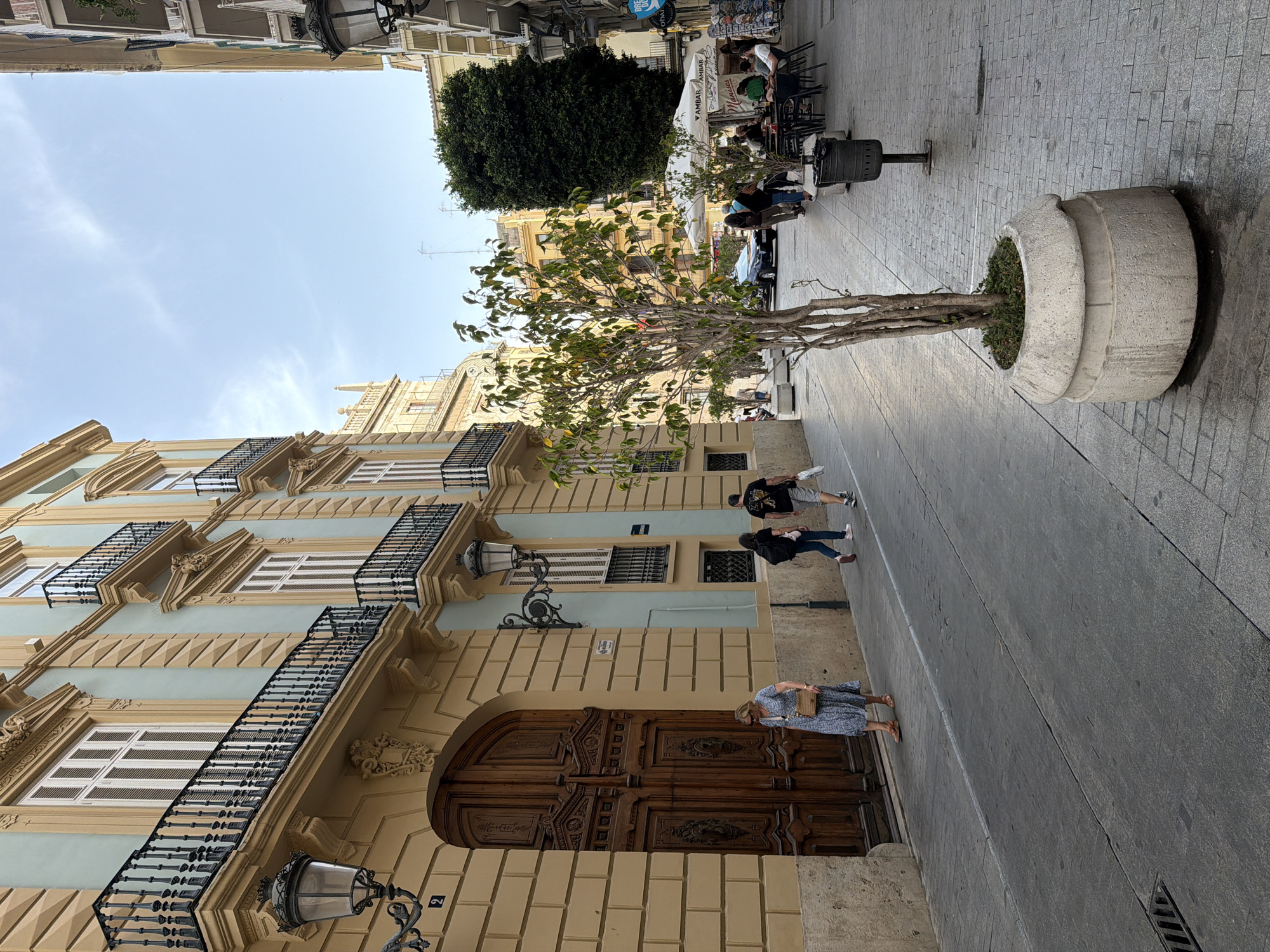 An outdoor shot shows a narrow city street in what appears to be a European location. The street is paved with light gray cobblestones. On the left side of the street, a building features a light yellow facade with ornate architectural details, including balconies with black wrought-iron railings. The building also has a large, intricately carved wooden double door. On the right side of the street, there are more buildings, some with balconies and one with a green tree growing in a large concrete planter. Further down the street, there is an outdoor cafe with tables and chairs set up under umbrellas. Several people are visible walking along the street and sitting at the cafe. The sky is blue with a few wispy white clouds. The lighting suggests it is daytime, possibly late afternoon or early morning, with the sun casting shadows on the street.