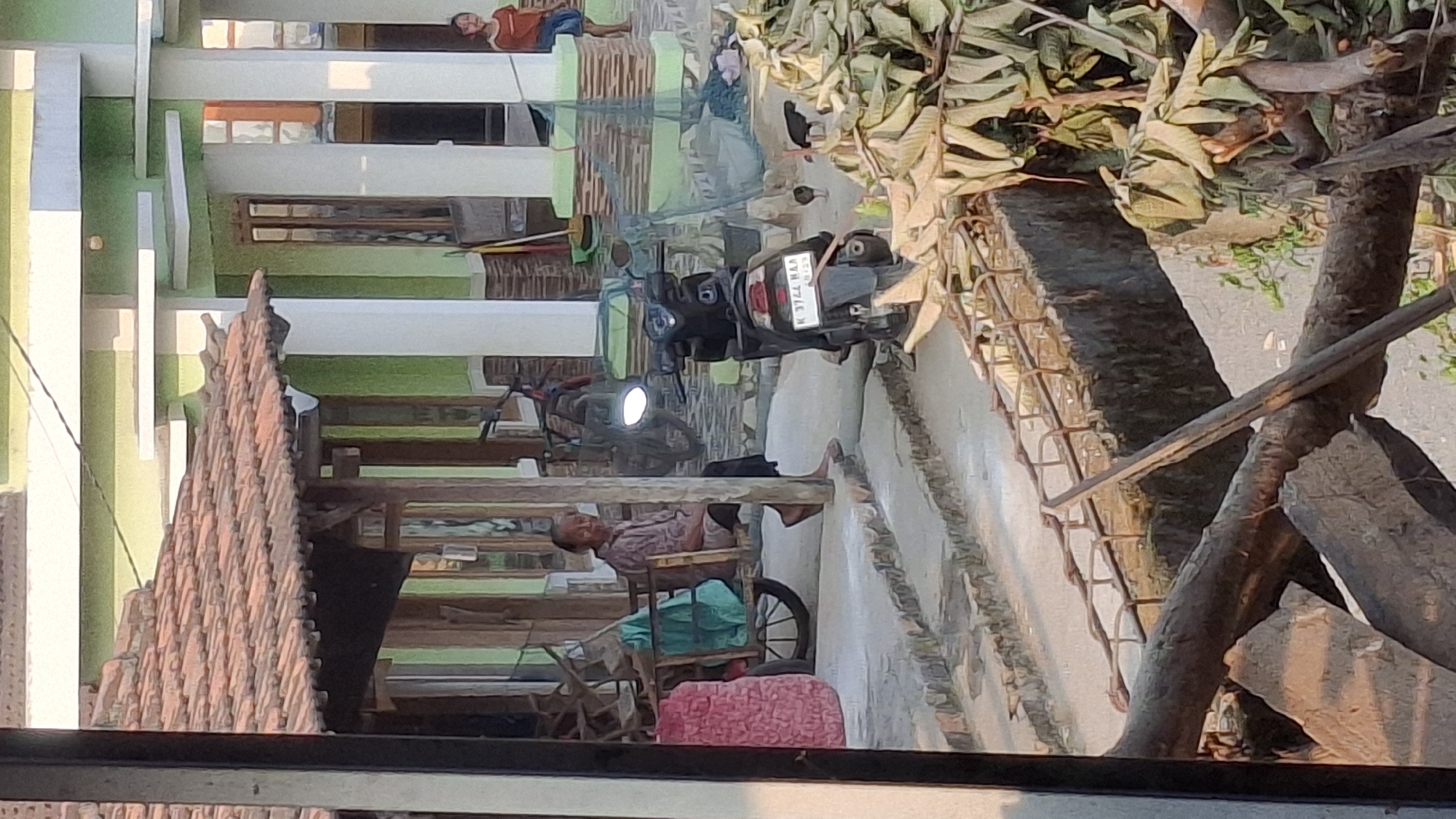 The image depicts a scene in Pucakwangi, Indonesia, showing a traditional outdoor seating area with a tiled roof. An elderly individual is seated on a wooden cart, with a green cloth draped over it. To their right, a black motorcycle is parked. In the background, a building with white pillars and green walls is visible. Further to the right, a child in an orange shirt stands near the building. The foreground of the image features a construction or repair site with exposed rebar and fallen branches. Several chickens are scattered around the motorcycle and a pile of dried leaves. The lighting suggests it is daytime, likely late afternoon due to the warm tones.