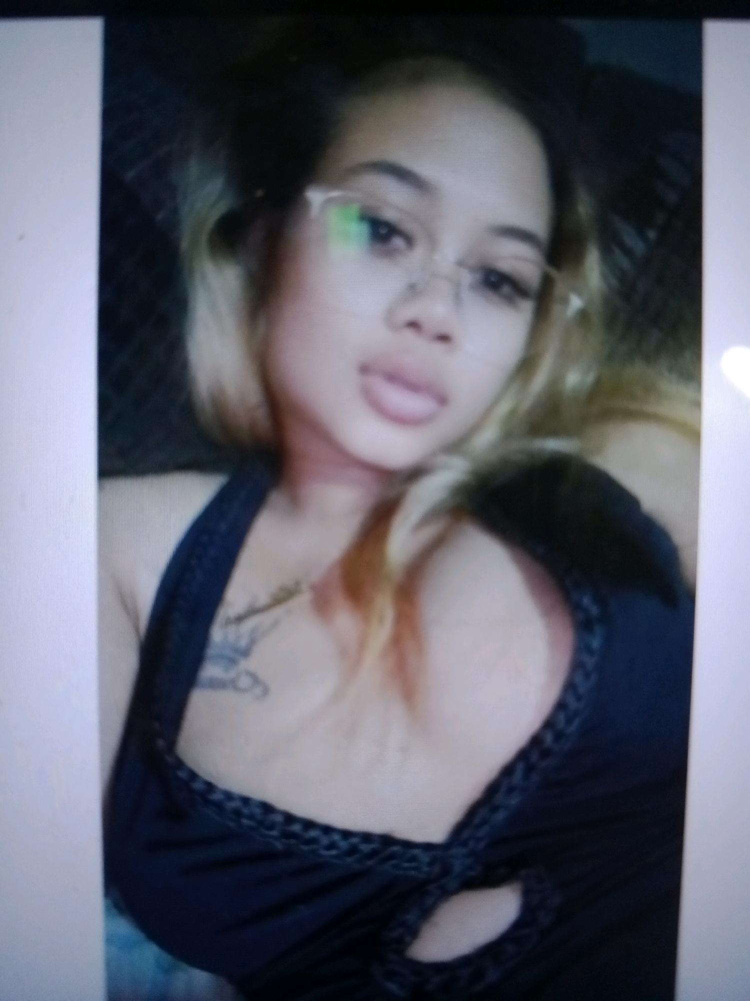 The provided image is a close-up, low-resolution photograph of a young woman. She has light-colored hair and is wearing glasses and a dark-colored top. A tattoo is visible on her chest. The background is dark and appears to be out of focus, suggesting a dimly lit indoor environment. There is no discernible information in the image that would indicate a specific location in Noord, Suriname.