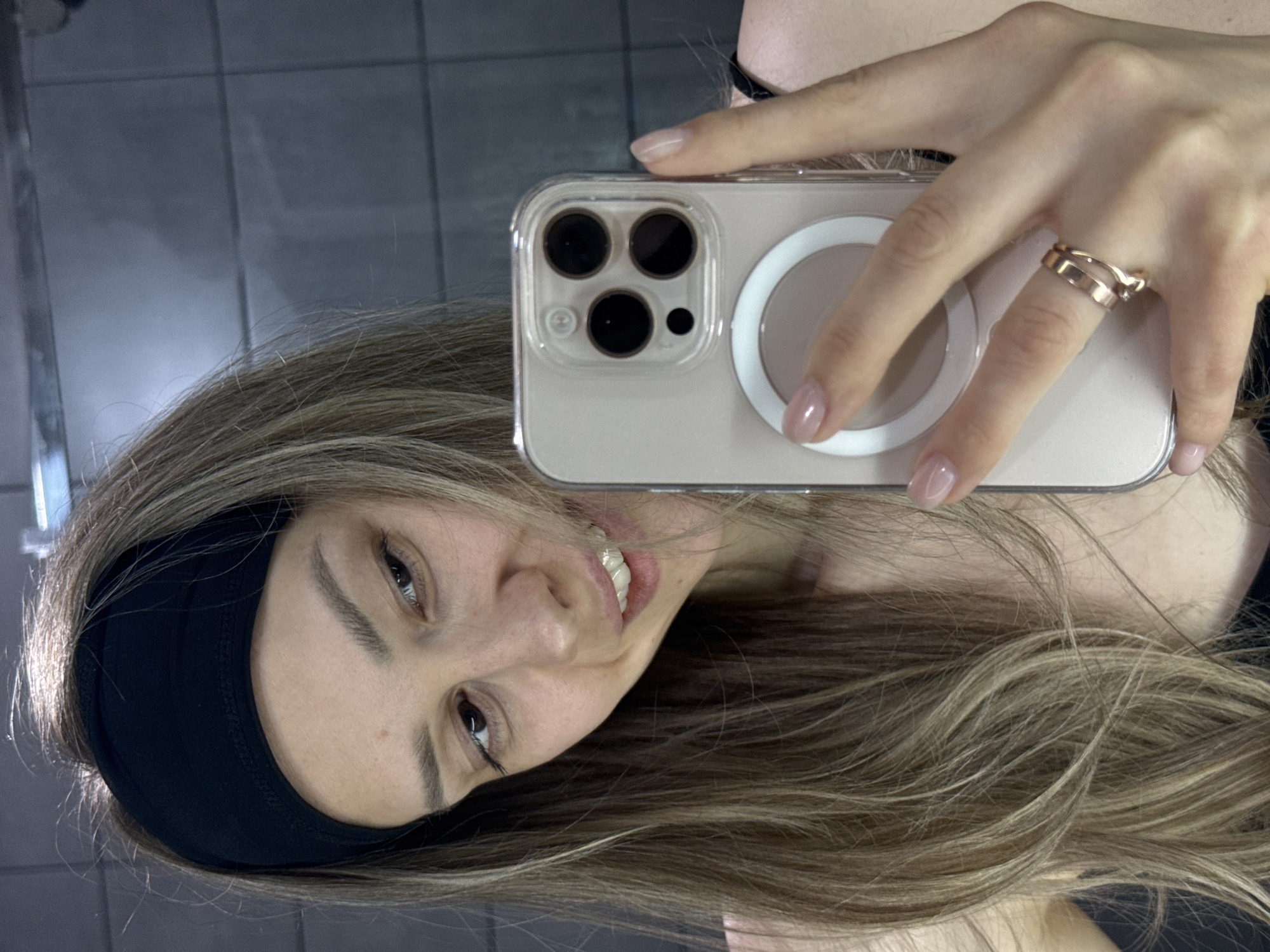 A woman is taking a selfie with her phone. She is wearing a black headband and a ring on her finger. The background appears to be a tiled wall, possibly in a bathroom or similar space. The photo was not taken in the city of Chaikovsky, Russia.