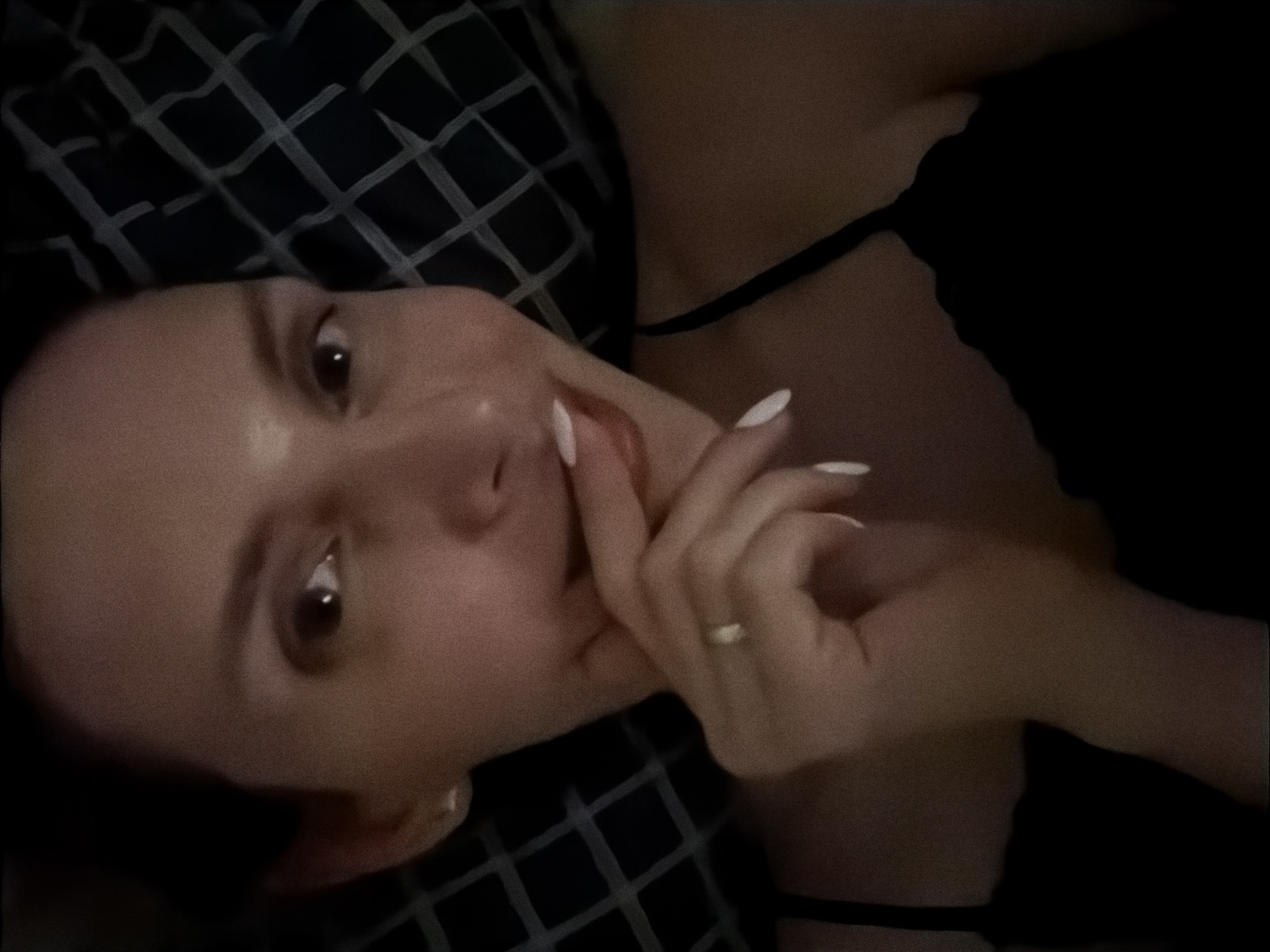 A woman lies on a surface with a dark blue and black square patterned cover. She is wearing a black top with thin straps. She is holding her hand to her face, with a ring on her ring finger. The location is unknown.