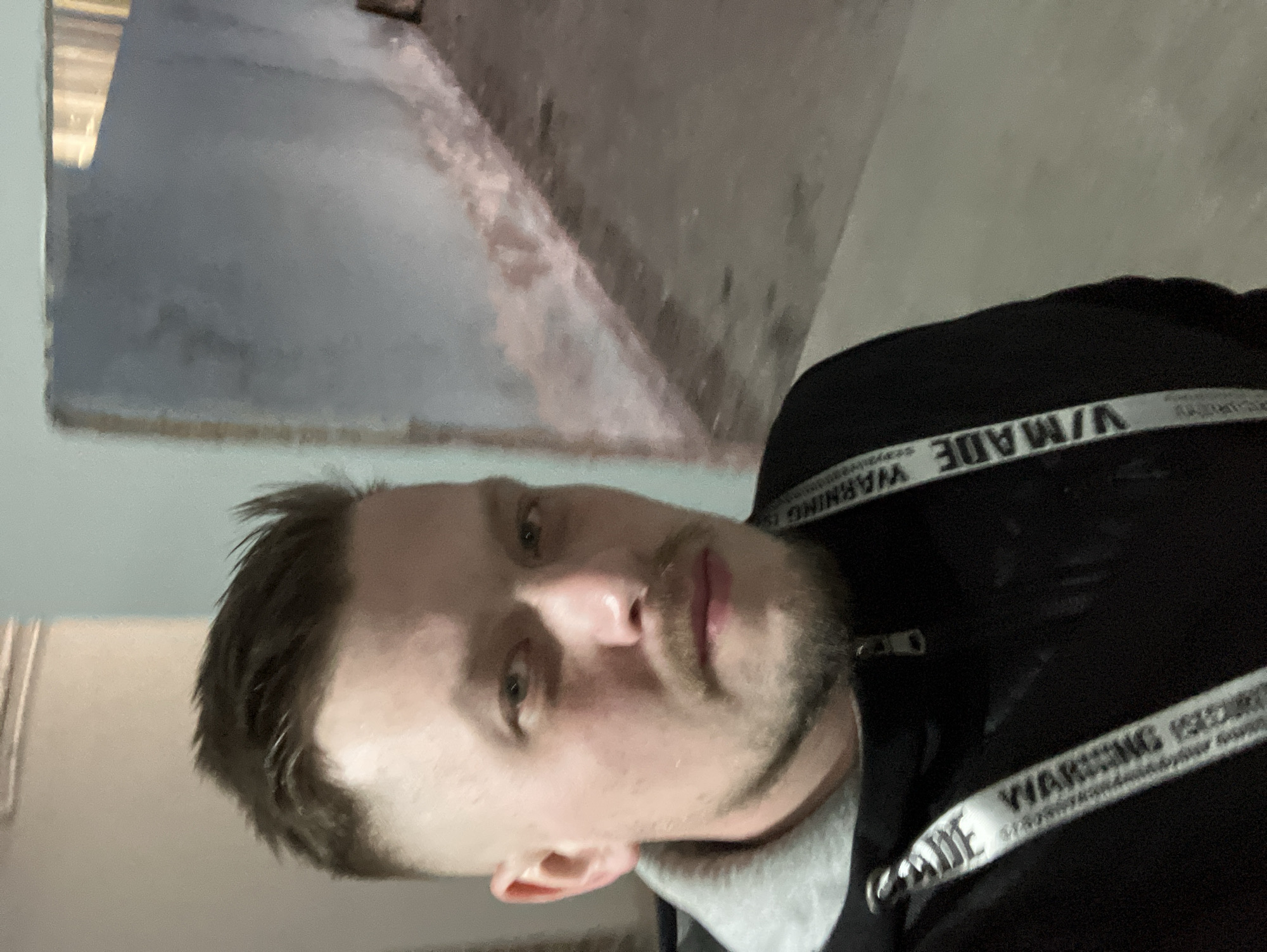A man is visible in the foreground. He has short brown hair, light skin, and stubble on his face. He is wearing a black jacket with white stripes that have the word "WARNING" printed on them. In the blurred background, there's a gray wall, possibly concrete, showing signs of wear or damage. The image is rotated, and the location is not discernible as Noviy, Russia or any specific place from visual information.