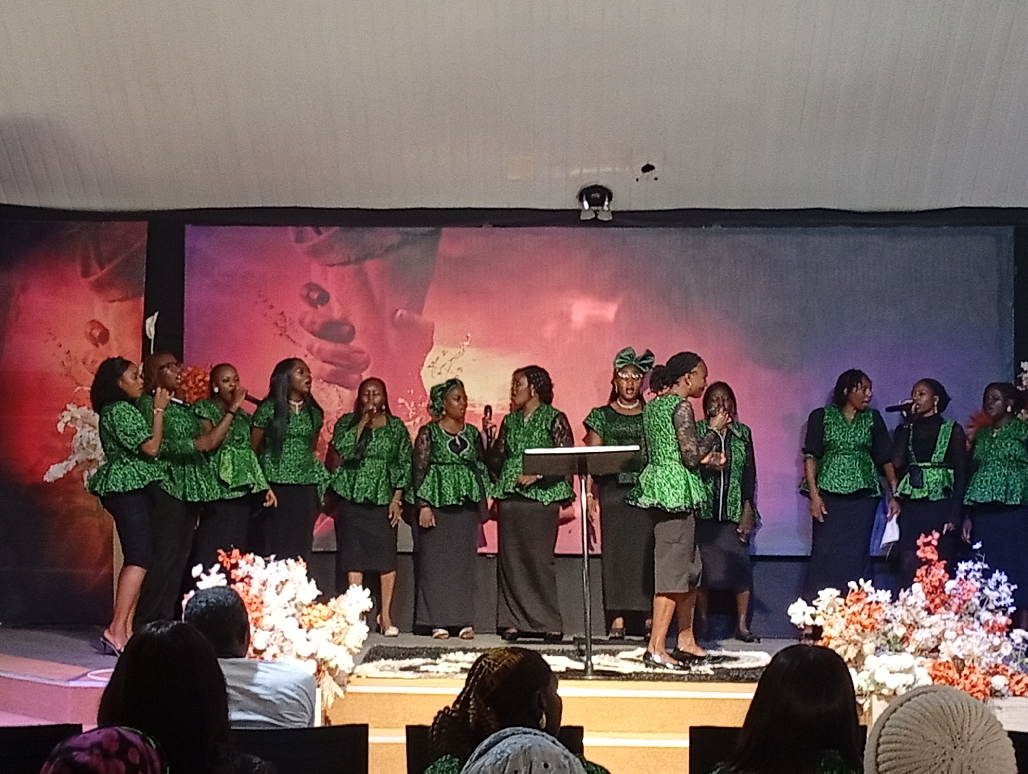 A group of approximately twelve women, dressed in matching green tops with black skirts or slacks, are standing on a stage in front of a large screen displaying a blurred, reddish-pink abstract image. The women appear to be singing, with some holding microphones. Several floral arrangements with white, orange, and pink flowers are placed around the stage. The stage is illuminated, and there is a black podium to the right of center. In the foreground, the backs of several audience members are visible, seated in chairs. The overall setting suggests a performance or religious gathering. This image was taken in Jos, Nigeria.