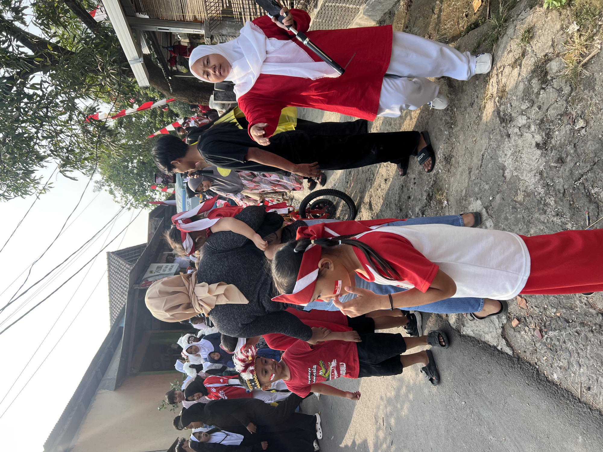 A group of people, including children, are gathered on a street in Klapanunggal, Indonesia, likely for a parade or celebration. Many are wearing red and white, the colors of the Indonesian flag, and some children have face paint resembling the flag. A young girl in the foreground, wearing a red and white outfit, is touching her face. To her right, a woman in a white hijab and red outfit is holding a long, dark object, possibly a flag pole or a decorative item. In the background, buildings line the street, and Indonesian flags are visible, some hanging from trees. The ground is paved with asphalt and some rougher patches. The lighting suggests it is daytime.
