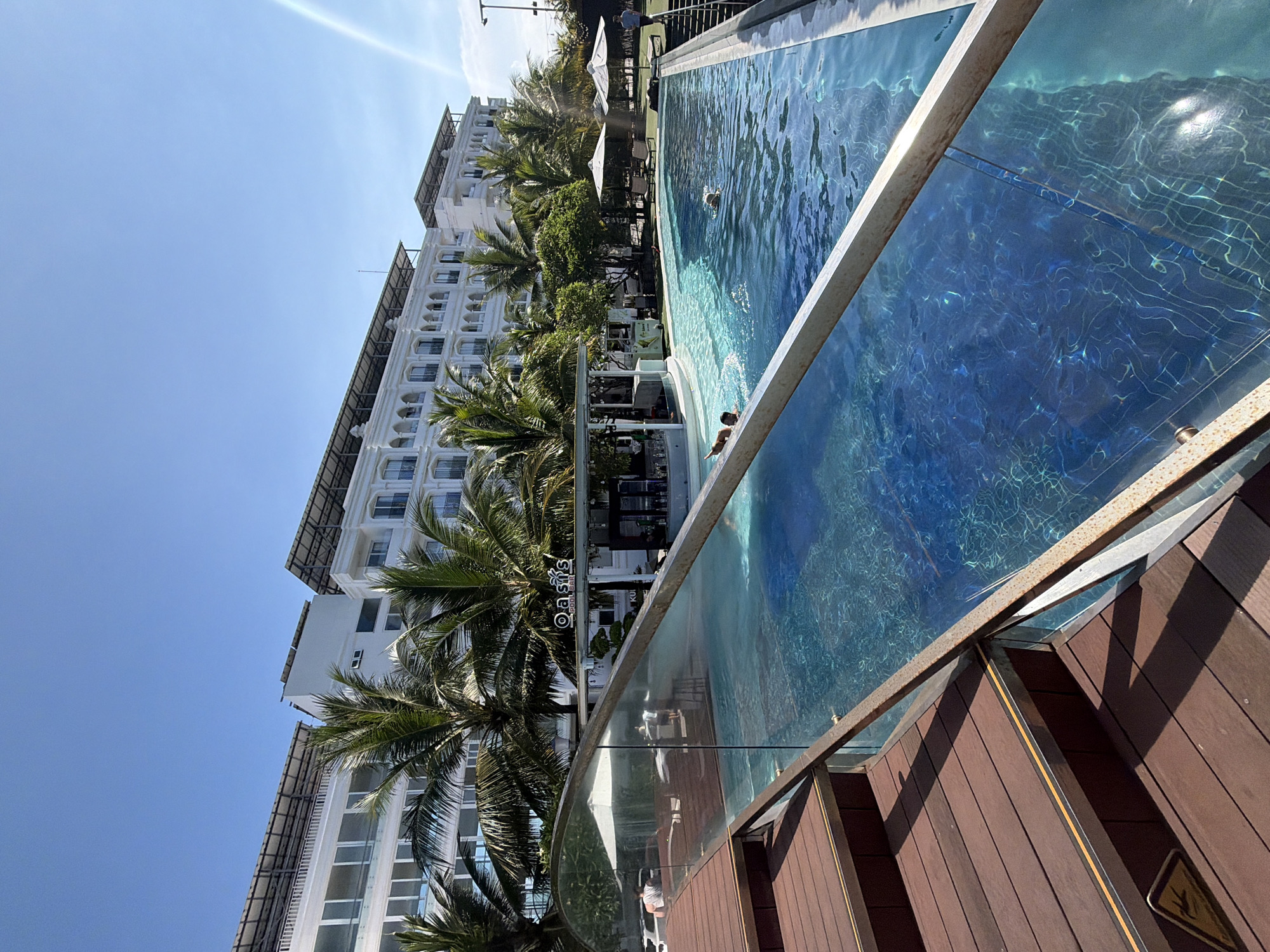 A sunlit exterior shot captures a resort-style swimming pool with a hotel building in the background. The hotel is a multi-story white structure with arched windows and balconies, topped with solar panels. Palm trees are scattered around the property. The pool, a vibrant blue, features steps leading into it and a swim-up bar labeled "Oasis Pool Bar." Several individuals are visible in the pool, with two people swimming and one person partially submerged. In the foreground, wooden stairs with a metal railing and a glass partition lead down to the pool area. The sky is a clear, bright blue with some lens flare on the right side.