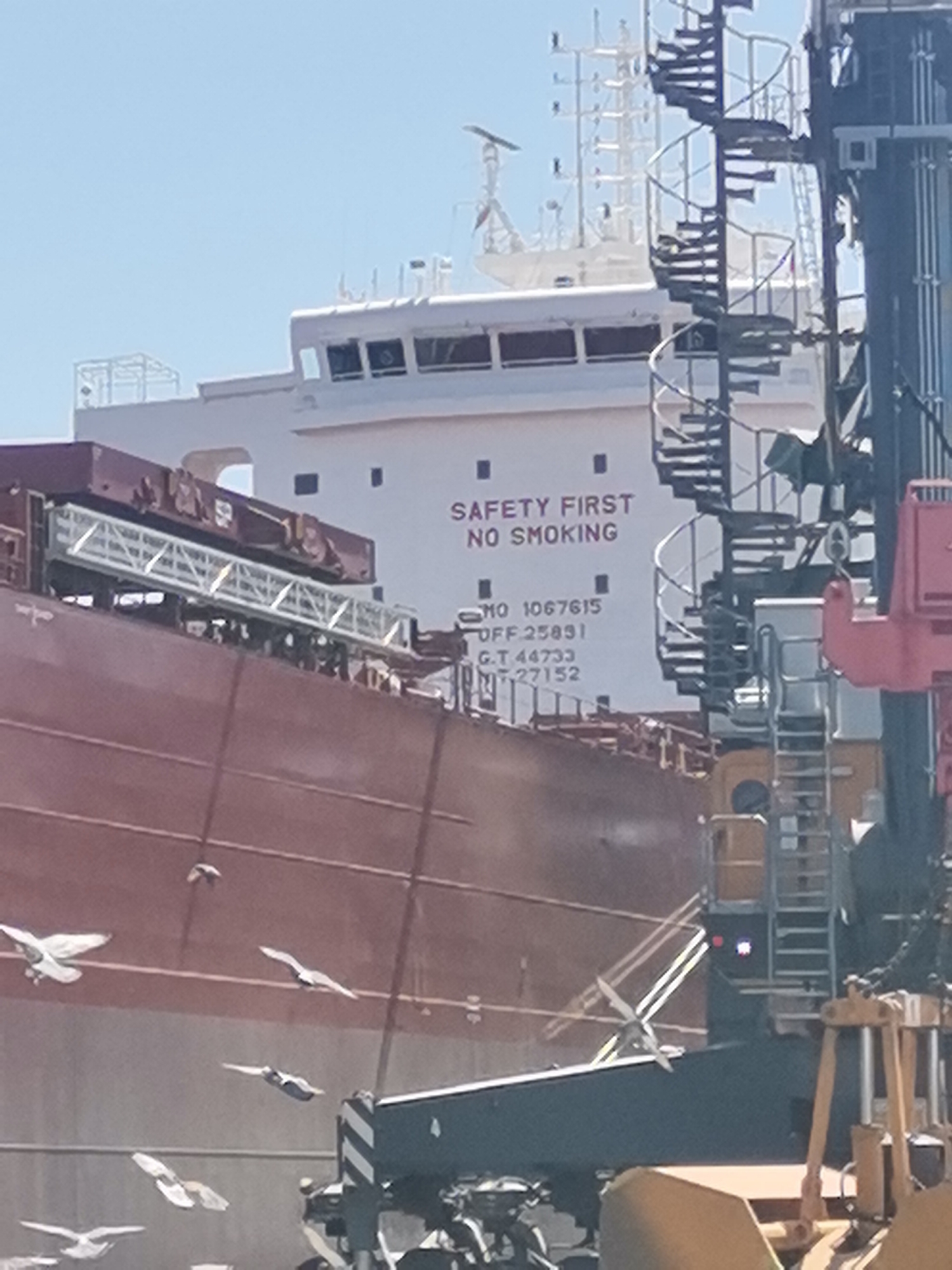 The image shows a large cargo ship docked in the port of Varna, Bulgaria. The ship is painted with a red hull and a white superstructure. In the center of the superstructure, "SAFETY FIRST NO SMOKING" is painted in bold, black letters. Directly below this message are registration numbers. A gangway extends from the ship to a dockside structure. To the right of the ship's superstructure, a spiral staircase and a crane are visible. Several seagulls are in the air in the foreground. The sky is clear and blue.
