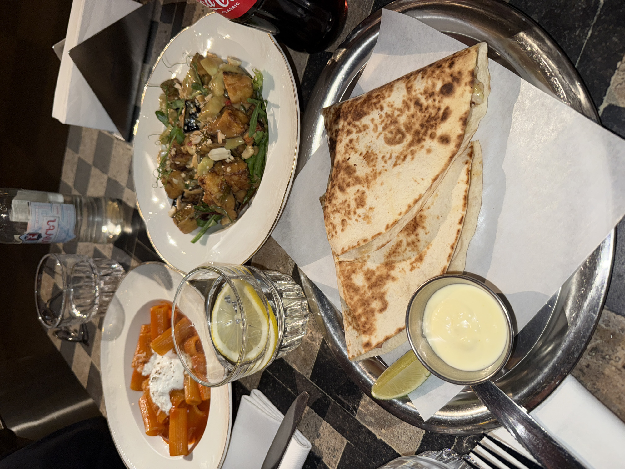 The image captures a table setting in what appears to be a restaurant, with a focus on various food and beverage items. The scene includes a plate of salad, another with pasta topped with a white sauce, and a quesadilla-like dish served on a metallic platter with a side of creamy sauce and a lime wedge. A glass of water with a lemon slice, a water bottle, and a Coca-Cola bottle are also present. Cutlery and napkins are neatly arranged alongside the dishes. The table has a patterned surface, possibly marble or stone. The location can be inferred as Yerevan, Armenia. The image suggests a meal setting in a restaurant environment.