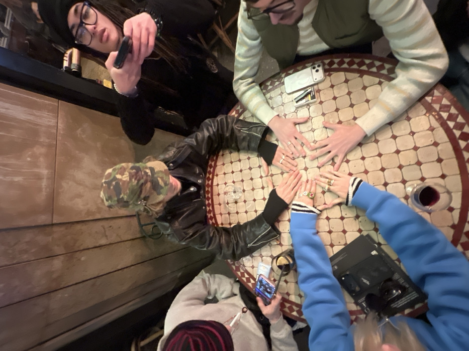 A group of five people are seated around a patterned, round table. Three people have their hands placed in the center, wearing rings. One person is wearing a camouflage hat and leather jacket, another a blue sweater, and the third a striped sweater. The other two people are on their phones. One wears a black beanie and glasses and the other has a red and black striped beanie. There are glasses of red liquid, phones, and black booklets scattered on the table. The table is set in what appears to be a bar or restaurant.