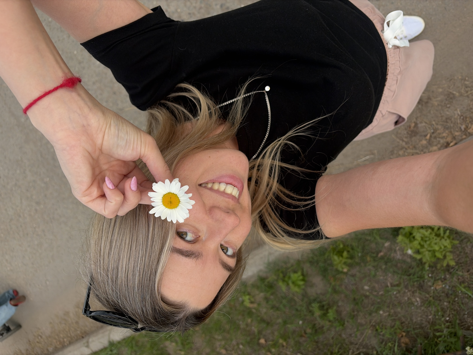 The media file is a selfie taken outdoors in Chaikovsky, Russia. The primary subject is a young woman with blonde hair, wearing a black t-shirt and pink shorts. She is holding a white daisy with a yellow center in her right hand, with the flower positioned in front of her eye. She has light-colored eyes and is smiling, with her teeth visible. She is also wearing a red string bracelet on her left wrist and a silver necklace. In the background, there is a grassy area with some dry leaves and dirt. The lighting suggests it is daytime, and the weather appears to be mild. The image is oriented upside down.