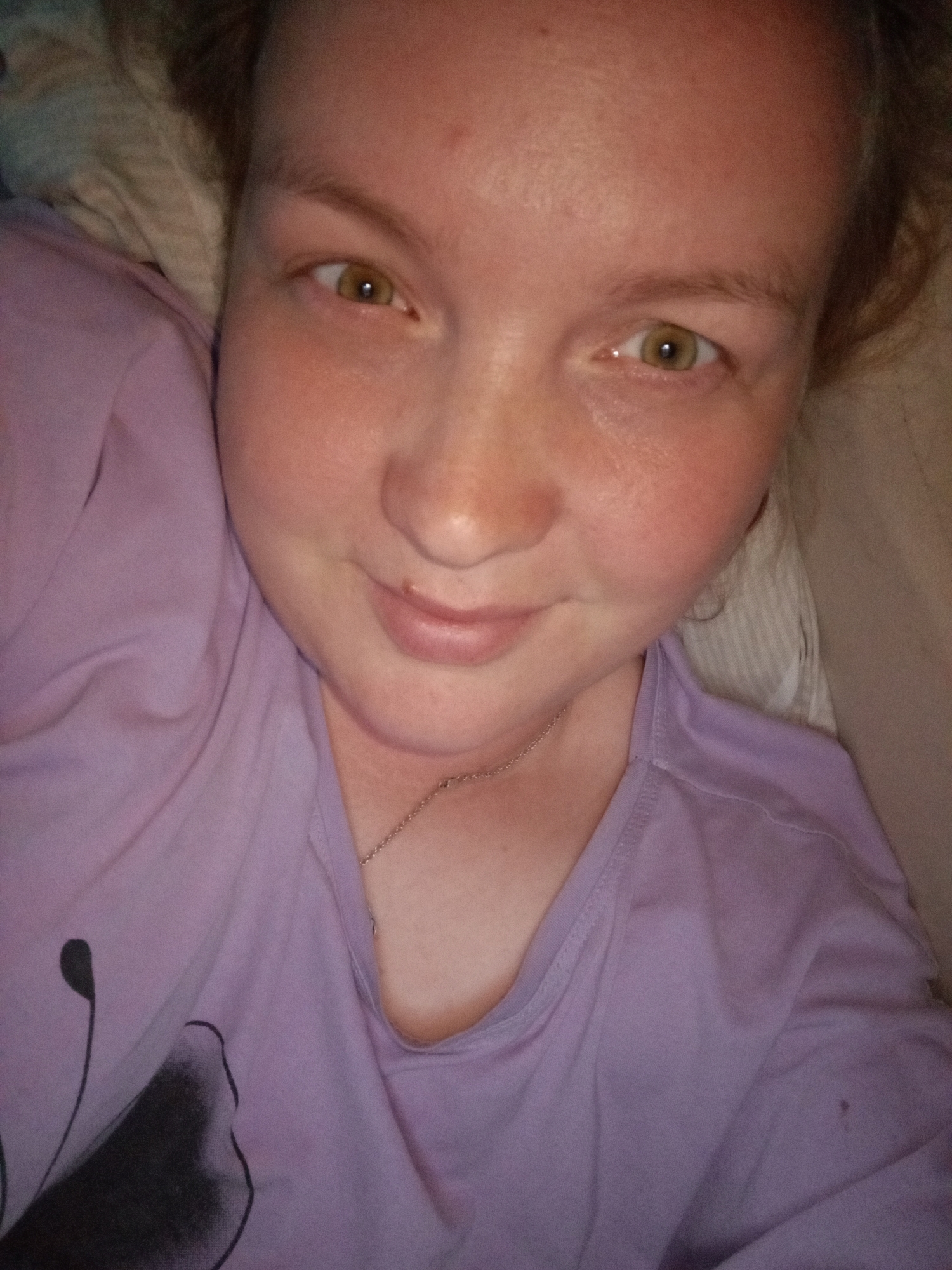 A person with fair skin and light brown hair is lying down, facing the camera. The person has brown eyes and a slight smile. They are wearing a lavender-colored shirt with a dark, abstract floral design on the left side. A thin silver chain necklace is visible around their neck. The person is lying on a white pillow. The location is Уральское, Russia.