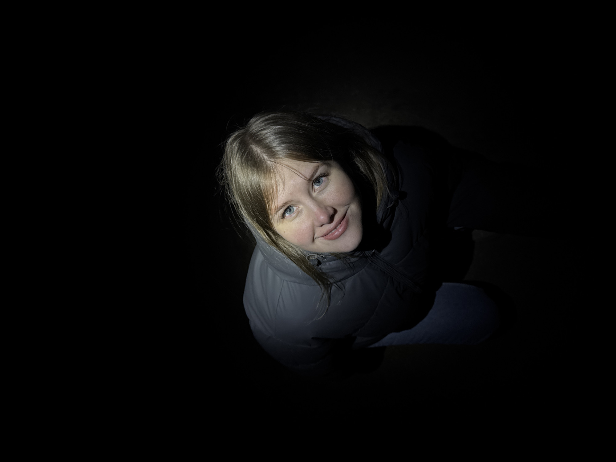 A young woman is seen from a low-angle perspective, lit from above against a completely dark background. She wears a gray puffy jacket with a zipper, and appears to be kneeling or crouching. The location is likely outdoors at night, but due to the heavy contrast, no details of the environment are visible. The image lacks sufficient information to determine the city, even if Горный, Russia, is the intended location.