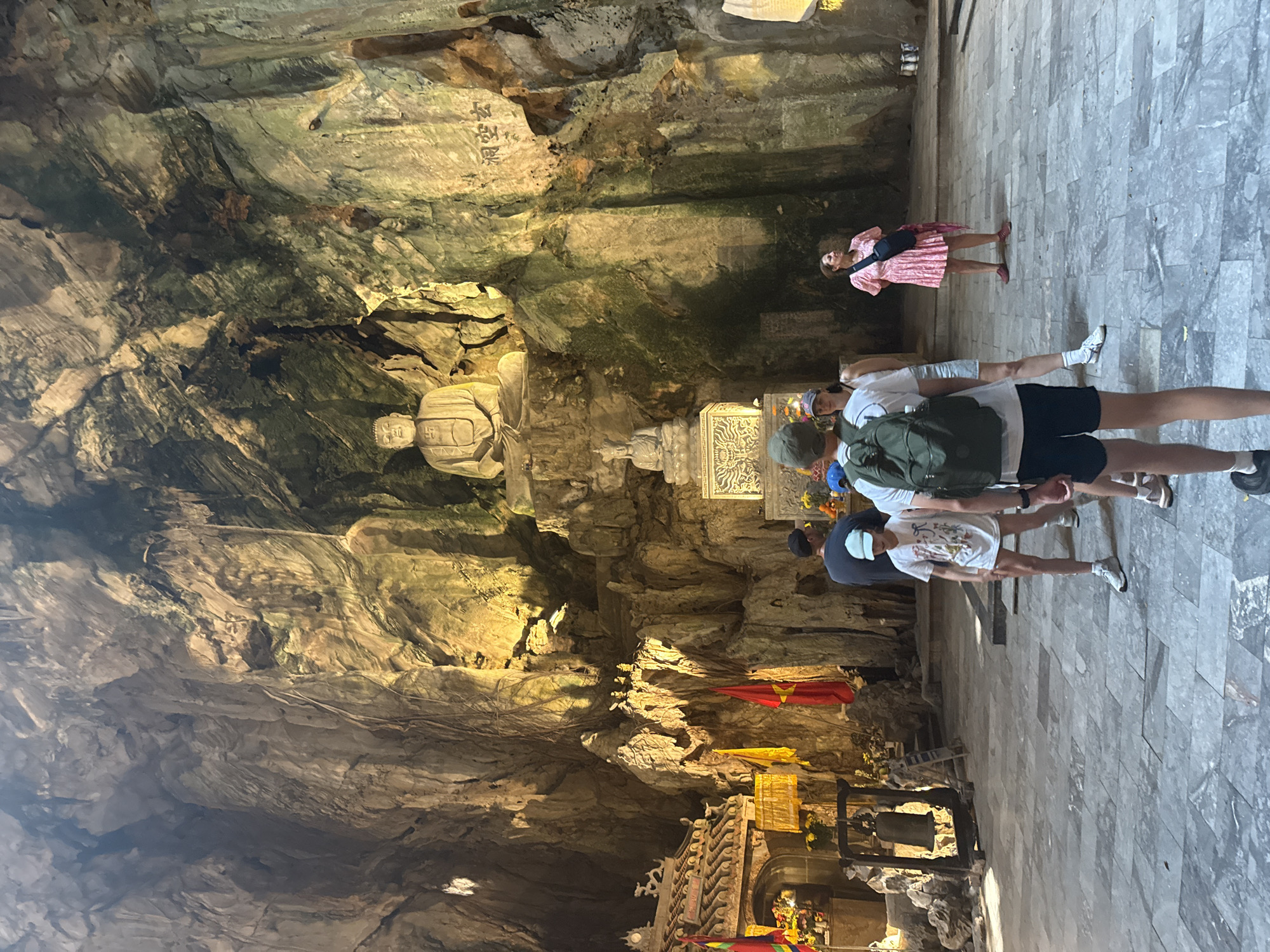 The image depicts a cavern within the Marble Mountains in Đà Nẵng, Vietnam. The cavern features natural rock formations, with a seated statue and script inscribed on the walls. Several tourists are present, including one woman standing and a group of three walking, facing away from the camera. Near the bottom of the frame, there are structures resembling altars or shrines.