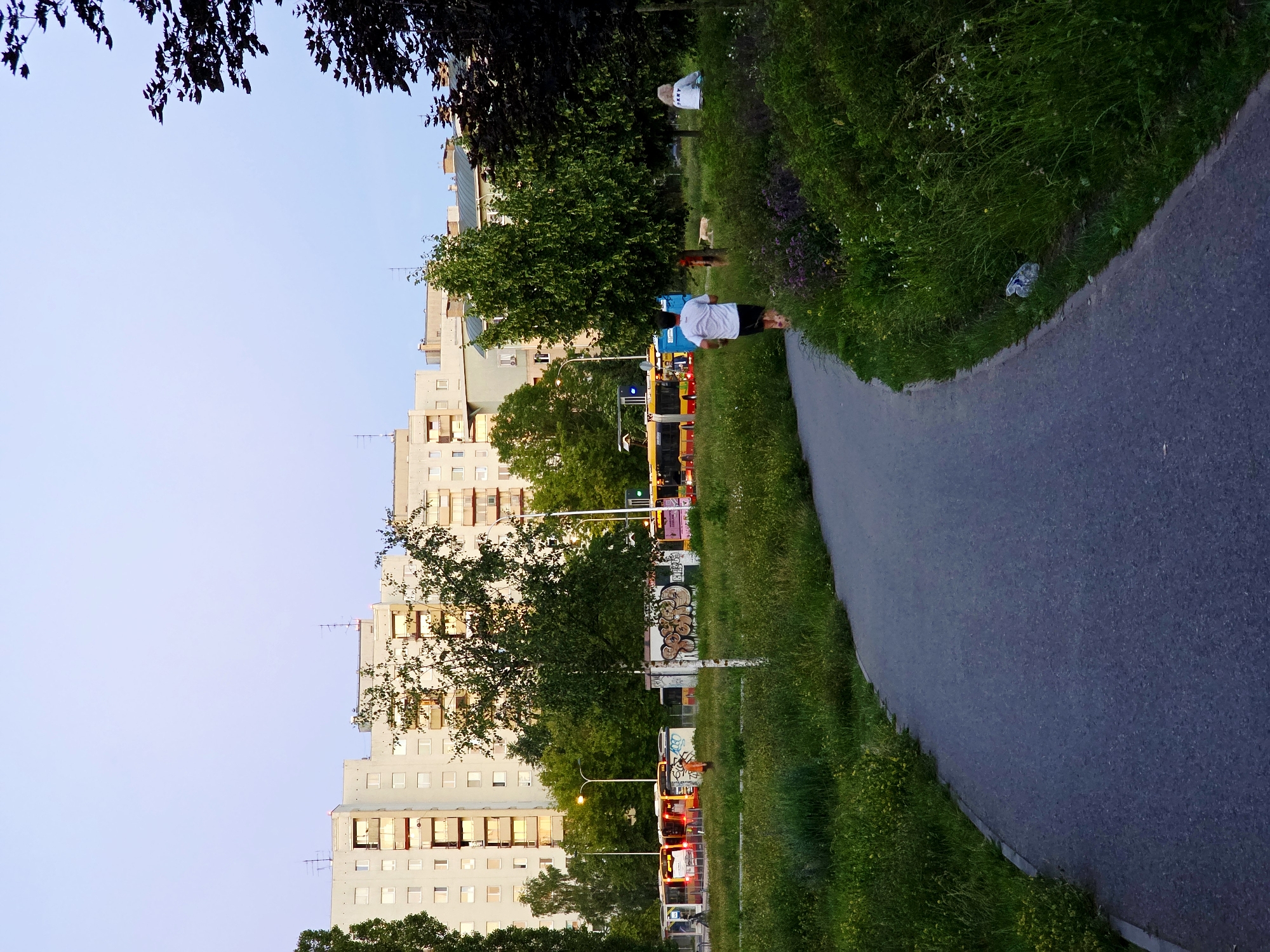 The image depicts a residential area in Warsaw, Poland, with tall apartment buildings in the background. A paved walkway curves through a grassy park where a man is jogging away from the camera. Several buses are parked along a street visible beyond the park. In the distance, two people are sitting in the grass, and a dog lies nearby. The sky is a pale blue, indicating either early morning or late evening. The scene suggests a typical urban environment with recreational spaces.