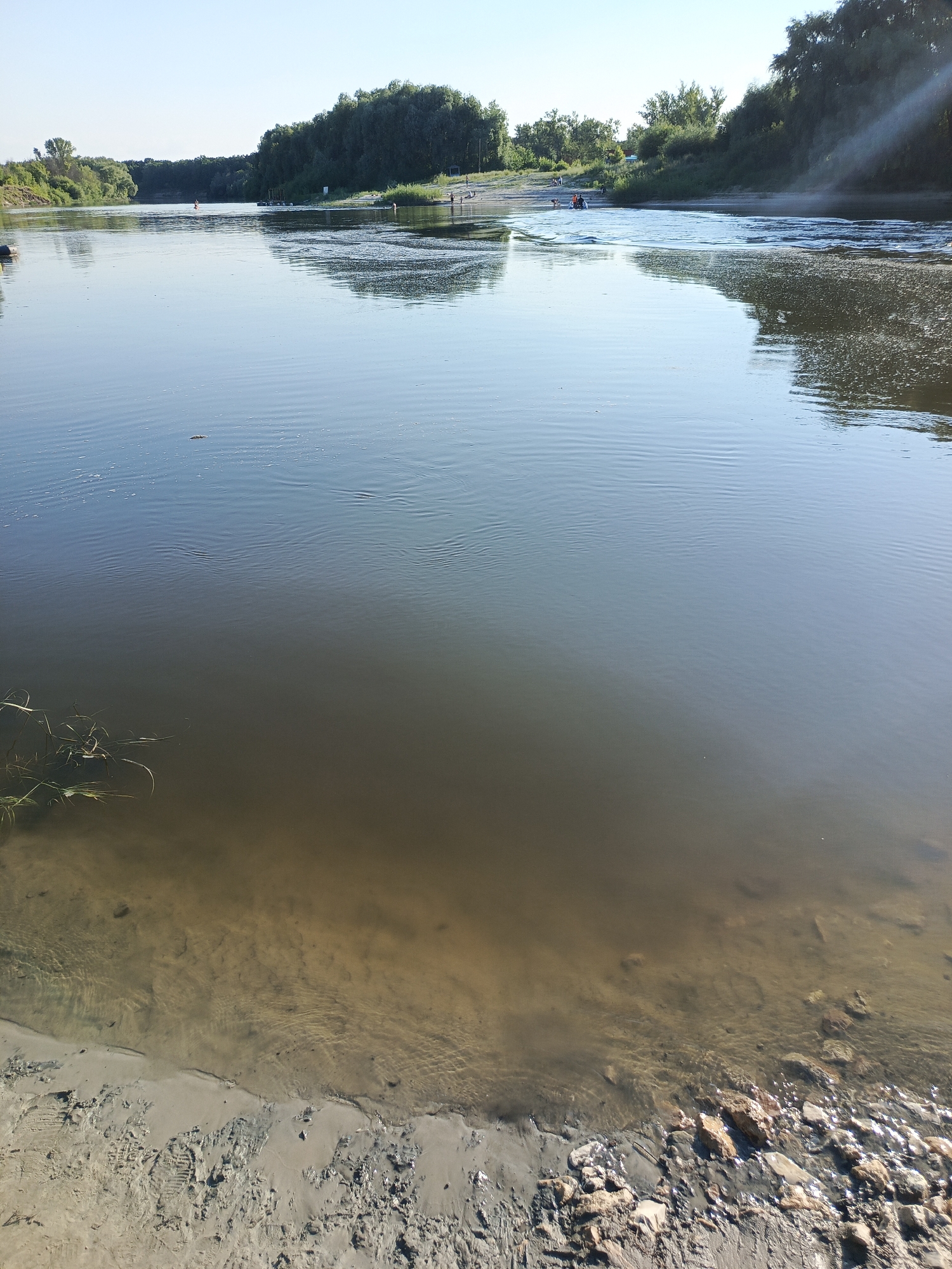 This is a photograph of a wide river on a clear, sunny day. The river takes up most of the frame, reflecting the bright blue sky. On the far bank, a dense line of green trees forms the horizon, with some smaller trees and vegetation closer to the water's edge. Several people are visible on the sandy bank on the right side of the image, appearing to be enjoying recreational activities, with some possibly wading or swimming. In the water, a small orange buoy is visible in the distance on the left, and further downriver, a small raft or boat with people on it is seen. The water in the foreground is murky, revealing a sandy bottom near the edge of the shore. The shore itself is composed of sand with some scattered rocks and debris. Ripples on the water's surface suggest a gentle current. The overall atmosphere is peaceful and suggests a warm, pleasant day, likely during the summer months. The sunlight casts a soft glow, indicating it is likely morning or late afternoon. The location is Uryupinsk, Russia. No text is visible in the image.