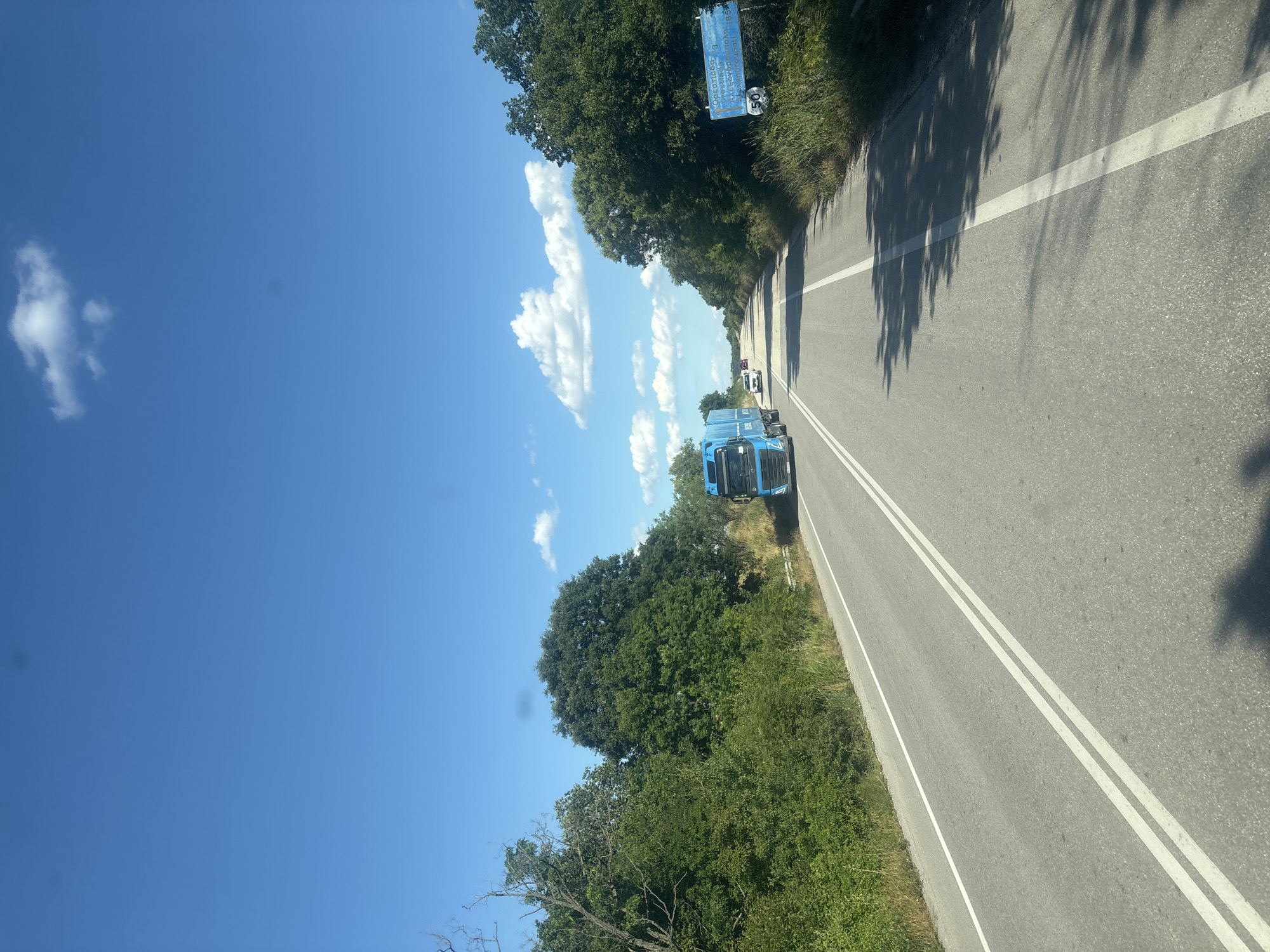 The image depicts a highway scene on a clear, sunny day with a bright blue sky and scattered white clouds. The road is asphalt with double white lines indicating two lanes in each direction. Lush green trees line both sides of the road, with dense foliage in some areas and sparser vegetation in others.
On the left side of the road, a blue truck with a white trailer is driving away from the camera. Further down the road, several other vehicles, including a white car, are visible. On the right side of the road, a blue and white sign with text in Greek and a speed limit of 50 km/h is posted. The sign appears to be a road sign indicating a town or region.
The overall scene suggests a journey through a rural or semi-rural area, possibly in Greece, given the Greek lettering on the sign. The environment is natural and appears to be in good condition, with well-maintained road markings and abundant vegetation. There are no people visible in the image, and the actions are limited to vehicles driving on the highway.