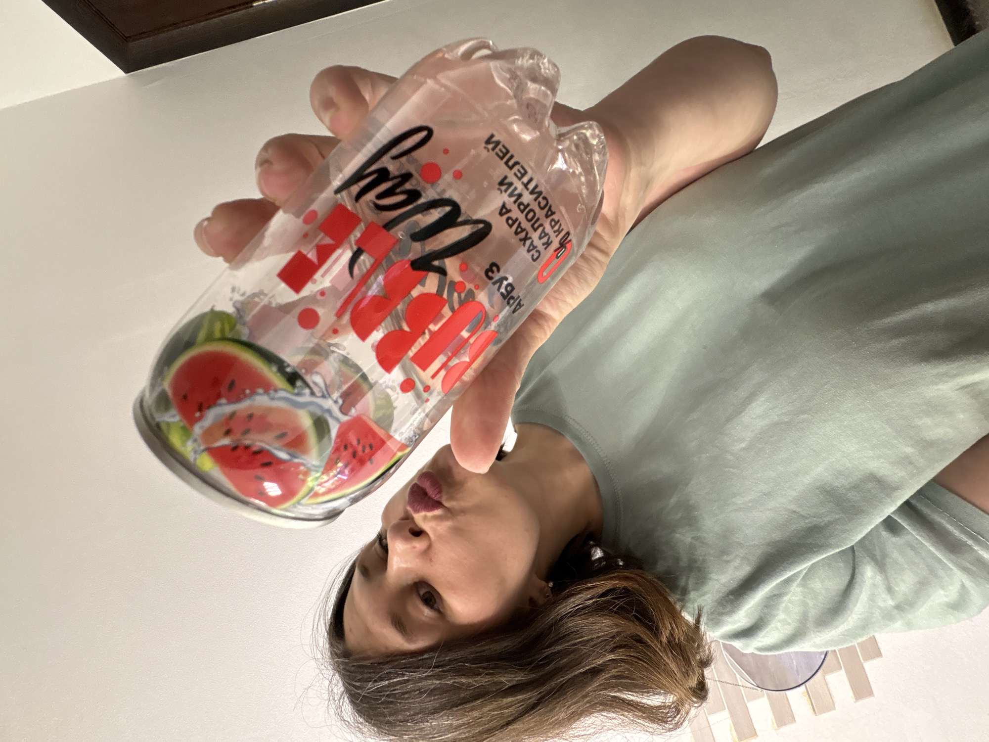 A woman with brown hair is holding a plastic bottle with a watermelon design and Russian text in her hand. The text on the bottle reads "Арбуз" (Watermelon) and mentions "0 сахара" (0 sugar). The photo was taken at a close-up angle, and the background is mostly white with some hints of a brown cabinet and possibly tiled walls, typical of an indoor setting. The woman is making a kissing face towards the bottle. The location is Chaikovsky, Russia.