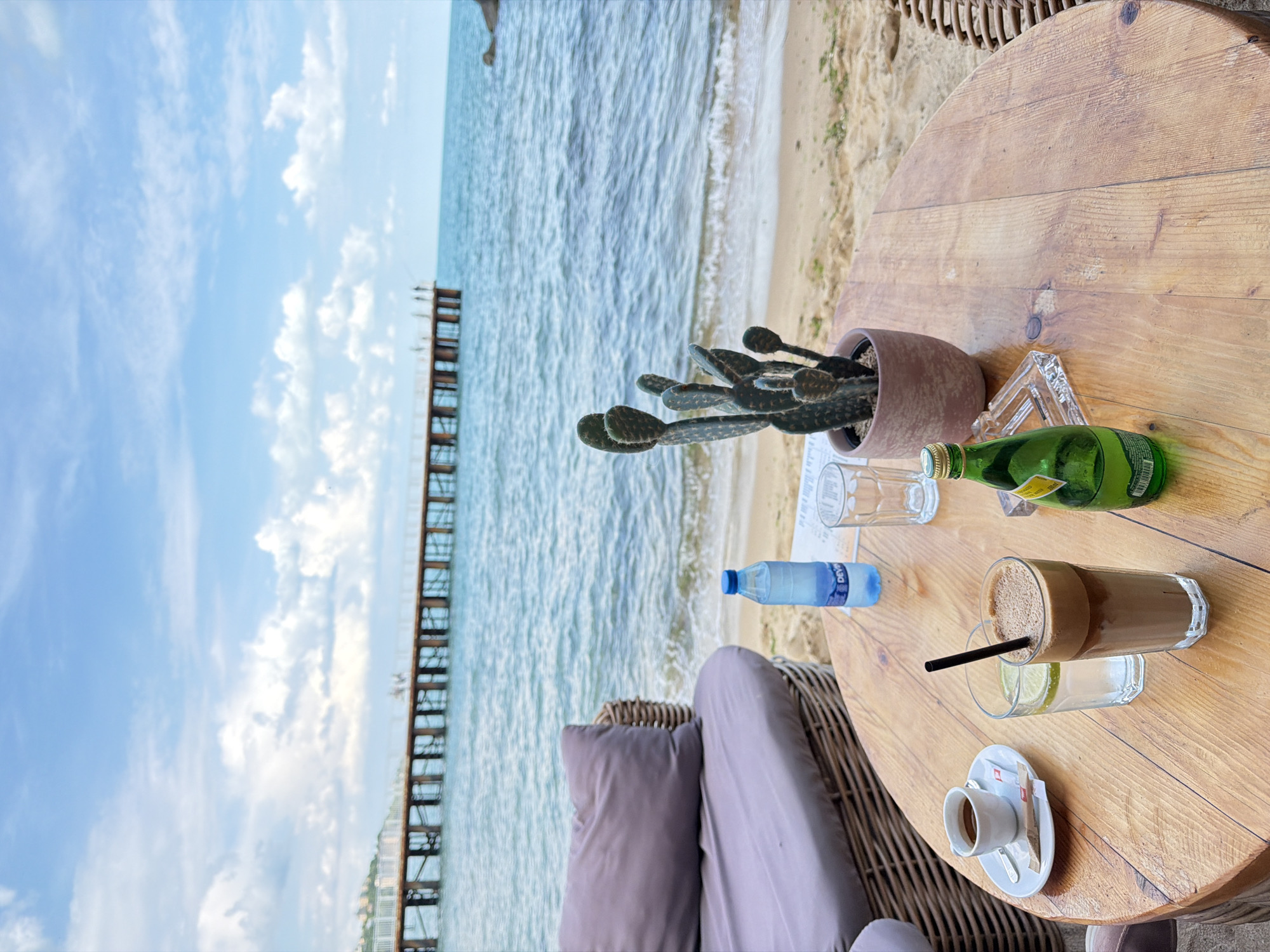 The scene is a table at a cafe overlooking the beach and sea in Varna, Bulgaria. On the wooden table, there's a cactus in a pot, two empty glasses, a bottle of water, a green bottle, an iced coffee drink, and an espresso on a saucer with a spoon and sugar packet. The view from the cafe includes a pier extending into the sea, the beach, and the sky with clouds. The cafe has a wicker-style chair or seating area.