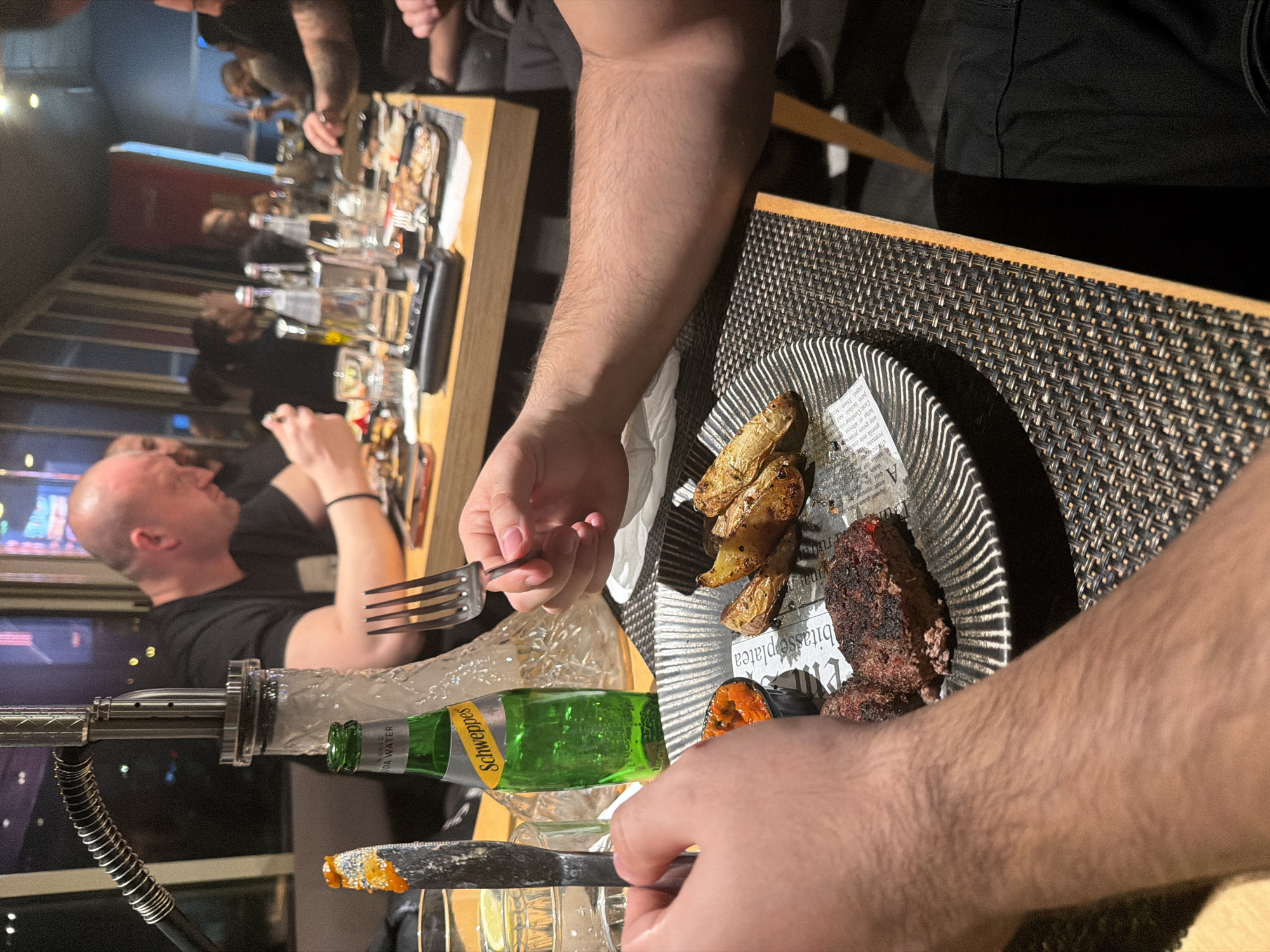 The photograph appears to be taken at a restaurant, likely in Plovdiv, Bulgaria, given the Cyrillic-infused script visible on the plate liner. A group of people is gathered around tables laden with food and drinks. The primary subject is a person holding a fork and a knife, poised to eat from a plate containing grilled potatoes and a piece of meat, possibly a burger patty. A bottle of Schweppes soda water is visible on the table, along with other glasses and bottles. The environment includes a window displaying a cityscape at night. The overall setting suggests a social dining experience in an urban setting.