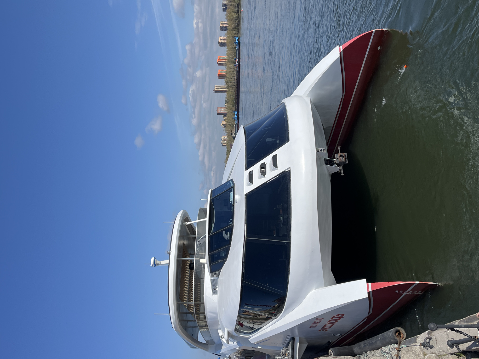 The image depicts a white and red catamaran-style passenger ship, named "EKOXOДЪ", docked alongside a concrete pier. The ship is facing to the right of the frame, with its bow partially submerged in the water. The water appears calm, with a slight greenish tint. In the background, across the river, there are buildings of various heights and colors, typical of a city skyline. Above, the sky is clear with a few scattered clouds. The overall scene suggests a sunny day at a riverfront location, presumably in Krasnoyarsk, Russia, given the name on the ship.