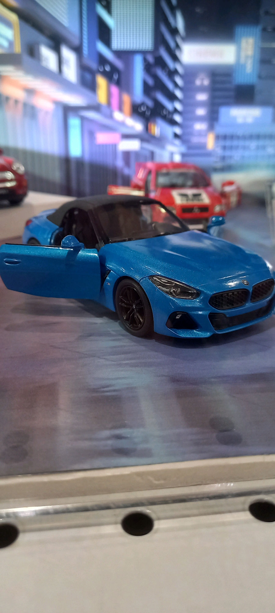 The image shows a blue convertible toy car with its driver's side door open, positioned on a surface that appears to be a diorama. In the background, there is a blurry depiction of a city street with buildings and lights. There are also other toy cars visible in the background, one red and one partially visible red car. The overall scene suggests a miniature display of a city environment, possibly a model or a toy store setting. There is no information in the image to suggest the location is Samarinda, Indonesia.