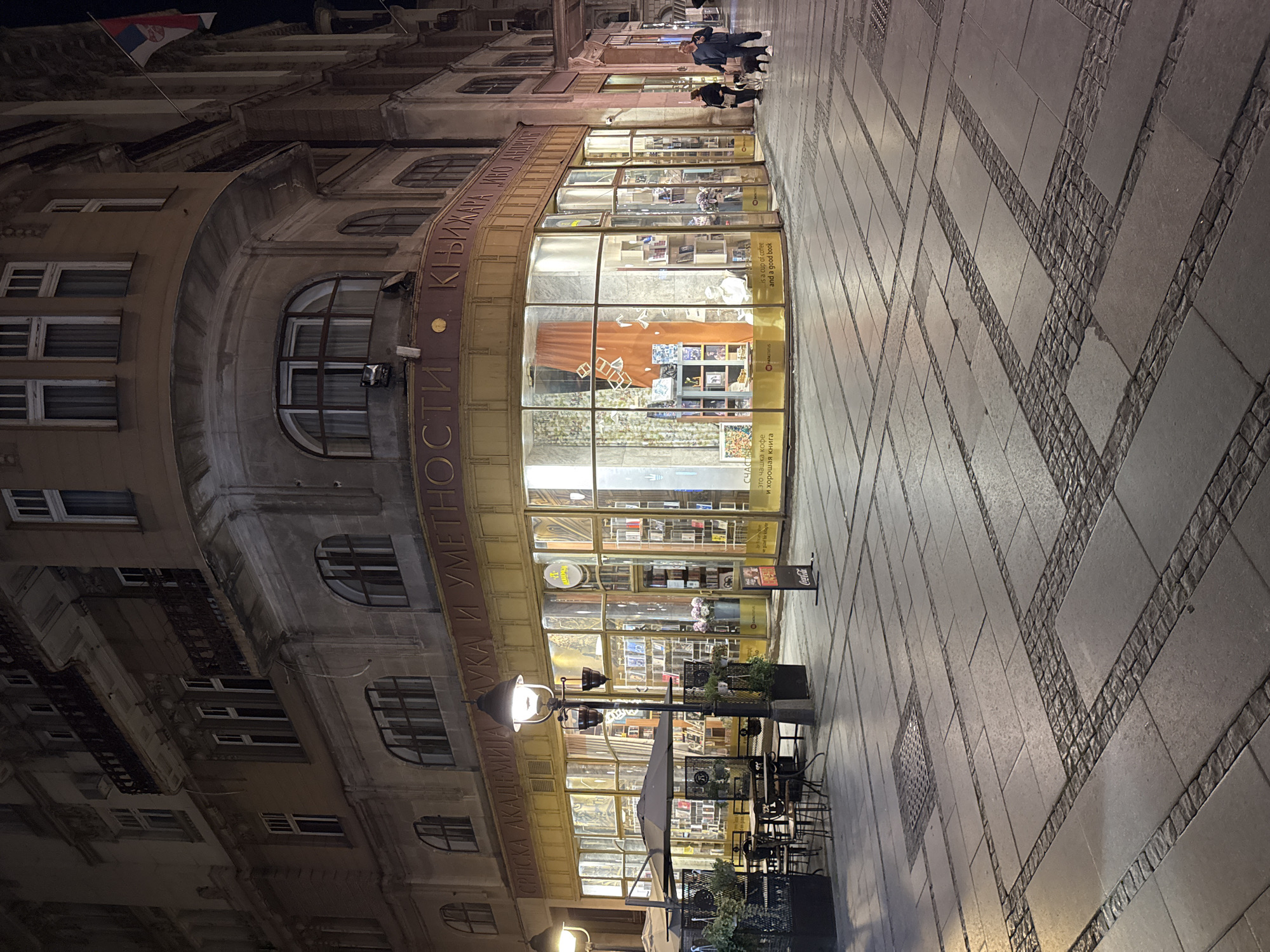 The image depicts a street scene at night in Belgrade, Serbia. The Srpska Akademija Nauka i Umetnosti (Serbian Academy of Sciences and Arts) bookstore is prominently featured. The store has a curved glass facade with shelves filled with books visible inside. A few people are walking on the sidewalk in front of the store. Tables and chairs from a cafe or outdoor seating area are also in front of the bookstore. A street lamp illuminates the area. The Serbian flag is visible atop the building. The sidewalk is paved with a pattern of tiles and cobblestones.