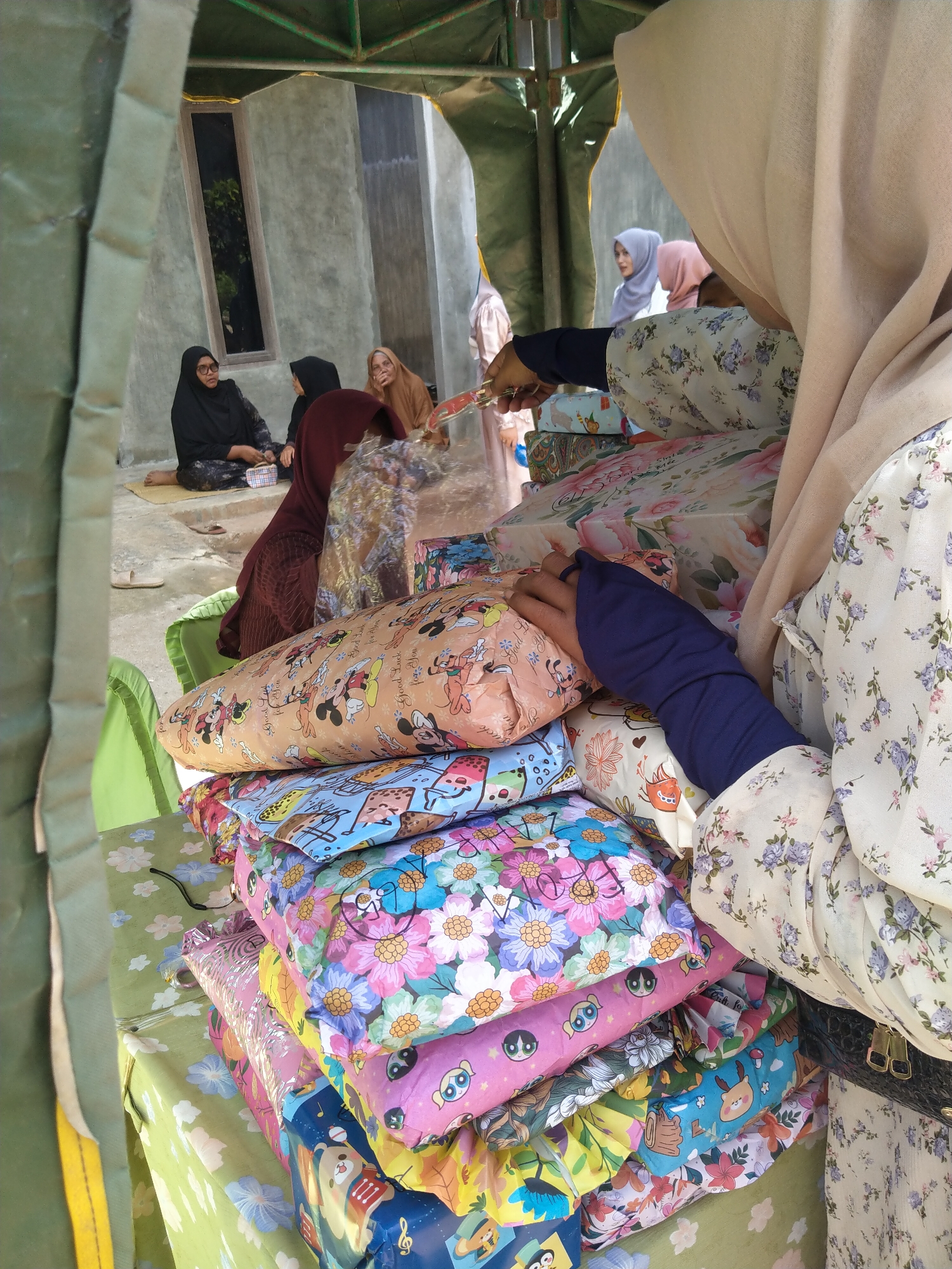 In Baratpayaitek, Indonesia, a group of women, some wearing hijabs, are gathered near a building with concrete walls and a window. In the foreground, several wrapped gifts are stacked on a table covered with a light green cloth adorned with floral patterns. The gifts are wrapped in brightly colored paper featuring various designs, including cartoon characters like Mickey Mouse and flowers. A woman, partially visible from behind, is handling one of the gifts, wearing a floral-patterned dress and a light-colored hijab. Another woman in the background, to the left of the gifts, is seated on the ground wearing a black hijab and glasses, holding a small patterned pouch. The scene appears to be an outdoor gathering, possibly a celebration or event, taking place under a green canopy.