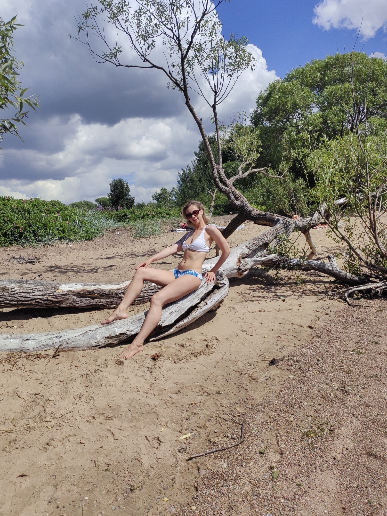 A blonde woman wearing a white bikini top and blue and white patterned bikini bottoms is sitting on a large fallen tree trunk on a sandy beach. She is wearing sunglasses and has her left arm resting on her leg. The tree trunk is weathered and grey. Behind her, there is lush green vegetation and trees, with a cloudy blue sky overhead. The scene appears to be a natural setting, likely a beach along the Neva River or Gulf of Finland, within the outskirts of Saint Petersburg, Russia, given the vegetation and climate. The sand is light brown and strewn with small pebbles and dried leaves.