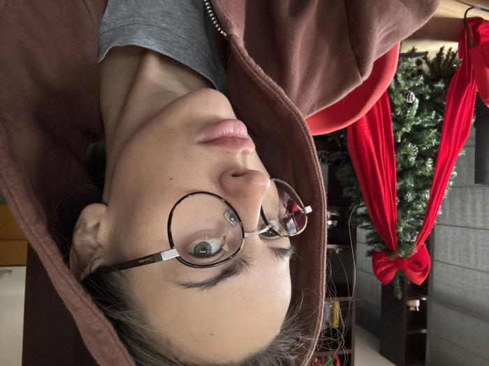 The image displays an adult female in a close-up, slightly off-center portrait shot taken indoors. She wears a muted reddish-brown hooded sweatshirt with the hood pulled up, revealing a grey zippered top underneath. Her face is towards the camera, with a neutral expression and slightly parted lips. She has fair skin, visible freckles, and light-colored eyes. She wears round-framed eyeglasses, featuring a tortoise-shell pattern on the lower rim and a silver-toned metal on the upper rim and bridge; the lenses reflect an indoor window scene. Her gaze is directed slightly to her left, towards the upper right of the frame. In the background, to the subject's left, an artificial green Christmas tree is visible. The tree is adorned with a large red fabric bow at its top and long, flowing red fabric ribbons draped vertically. Small pinecone ornaments are also present on the tree. Partially visible to the far left of the frame is a red chair or furniture item. Behind the Christmas tree, dark wooden shelving units are visible against a lighter wall. The specific location within the city of Warsaw, Poland, cannot be determined from the visual information provided in the image.