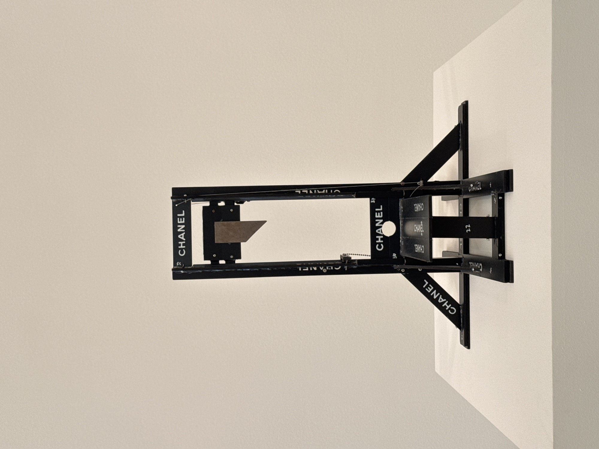 The image shows a black guillotine sculpture with "CHANEL" branding on various parts. The sculpture is positioned on a white pedestal against a plain, light-colored wall. The guillotine has a sharp blade, a wooden block at the base, and a chain mechanism. The "CHANEL" logo is visible on the top crossbeam, on vertical supports, and on the base. Small numbers are also present, such as "3/6", "37", and "22". The lighting suggests an indoor setting, possibly an art gallery or museum. The image is taken from a frontal, slightly low angle. There are no people or any other discernible objects or actions in the scene. The location in the city of Bilbao, Spain, is not specified in the image.