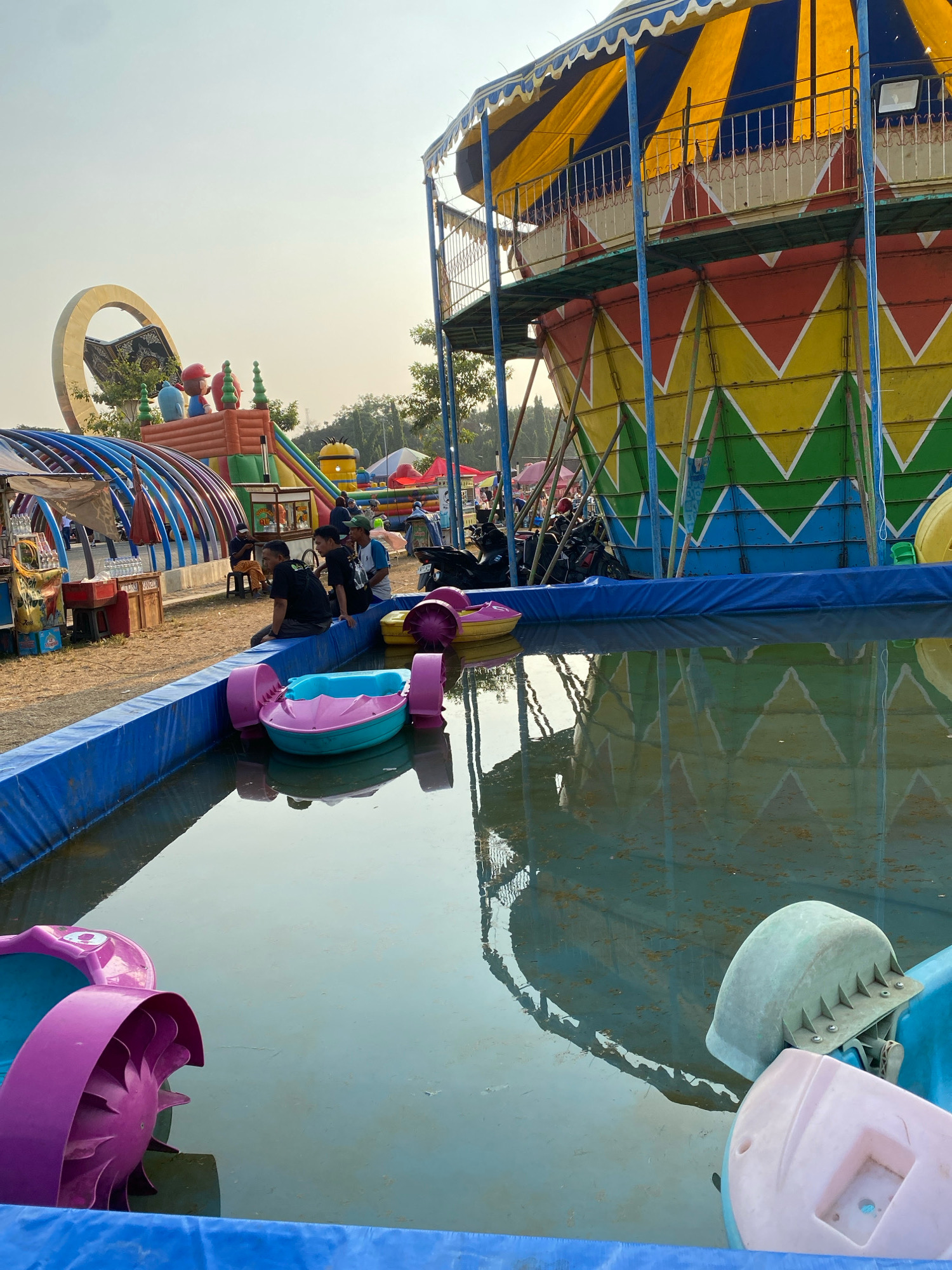 This is a medium-shot, outdoor photograph taken at a fairground in Nyamok, Indonesia. The scene depicts a temporary amusement park setup with various attractions. In the foreground, a small, blue-lined pool contains two pedal boats, one pink and turquoise and another pink. Several people are seated on the edge of the pool, facing the water. Behind the pool, various rides and attractions are visible. To the right, a colorful, circular ride structure with blue, yellow, and red diamond patterns dominates the background. To the left, a large, inflatable structure with cartoon characters is present, along with a curved, blue-arched walkway and a food stall. The overall environment suggests a lively, community gathering space with entertainment for families.