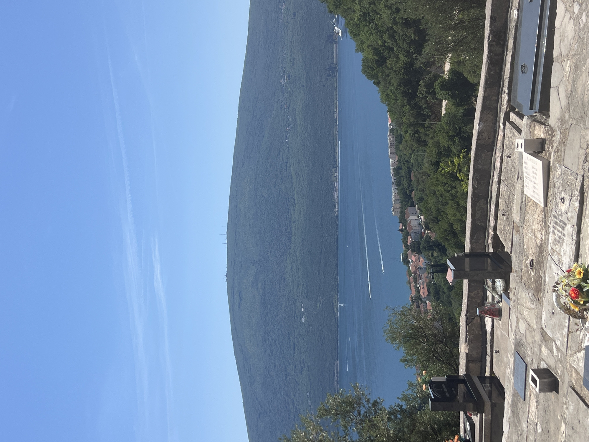 The image shows a view from what appears to be a cemetery or memorial site in Baošići, Montenegro. The scene overlooks the Bay of Kotor, with a large mountain partially obscuring the opposite shore. Several boats are visible on the water. The sky is clear and blue with contrails. In the foreground are weathered gravestones and memorial markers. Trees are positioned along the hillside. The city itself is visible along the waterline.