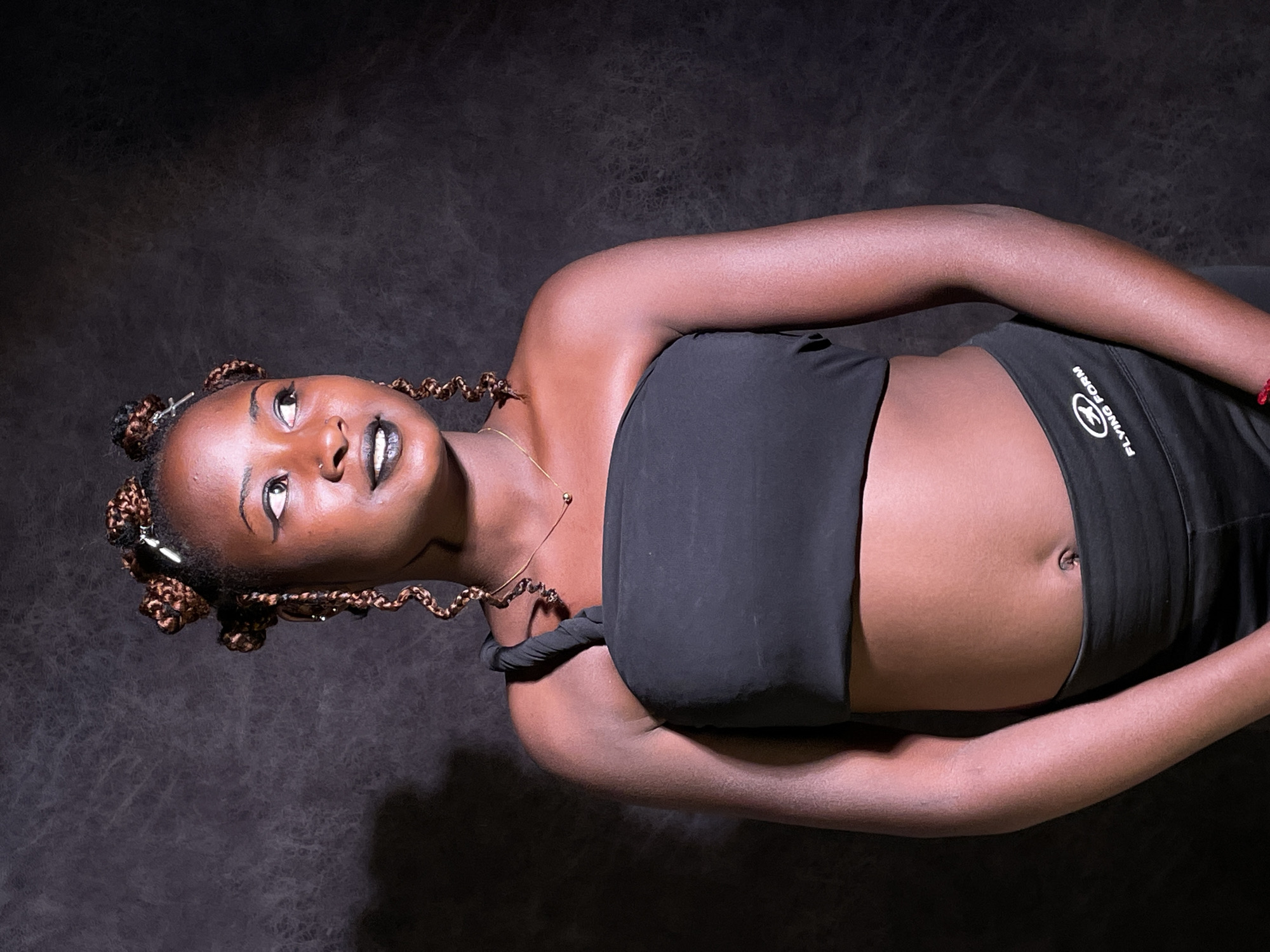 The image is an overhead portrait of a young Black woman lying on her back, looking upwards and slightly to her left. Her body is positioned diagonally across a dark, textured background.
She has dark skin and her hair is styled in multiple small, intricate braids gathered into decorative buns or knots, with some coiled tendrils. Her makeup includes prominent black eyeliner, light-colored eyeshadow, and dark lipstick. She wears a small nose ring and a delicate gold chain necklace with a small pendant.
The woman is dressed in a black strapless crop top, revealing her midriff, and black high-waisted leggings or athletic pants. On the hip of her leggings, there is a white circular logo and the text "FLYING FORM". A red bracelet is visible on her right wrist. Her arms are bent, with her hands resting on her abdomen. The lighting is dramatic, creating highlights on her skin and features against the deep, indistinct background. The expression on her face appears serene or contemplative as she gazes upwards.
The setting is an indoor space, possibly a studio, in Barakin Akawo, Nigeria. There are no other discernible objects or activities.