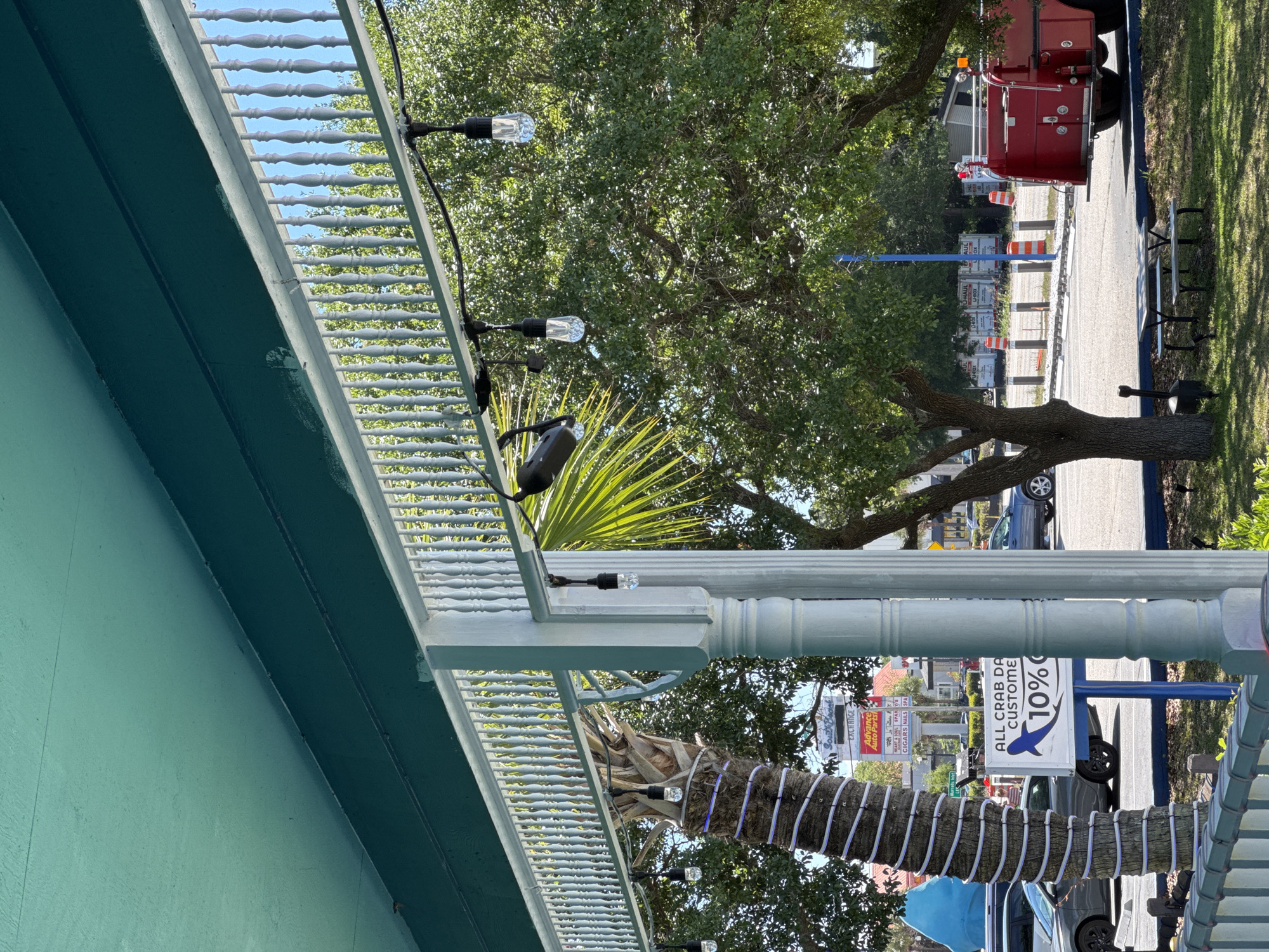 The image, taken in Garden City Beach, United States of America, shows an outdoor scene with elements of a building facade and a street view. The lower left portion features a turquoise-painted wall with a white balustrade along the top. A string of lights is draped along the balustrade.
Beyond the building facade, a street is visible with cars and a red fire engine parked on the right. A tree trunk wrapped with white lights is present in the foreground. A sign advertising "ALL CRAB DA CUSTOMER 10%" is also visible. The background includes other storefronts and businesses, and trees are interspersed throughout the scene.