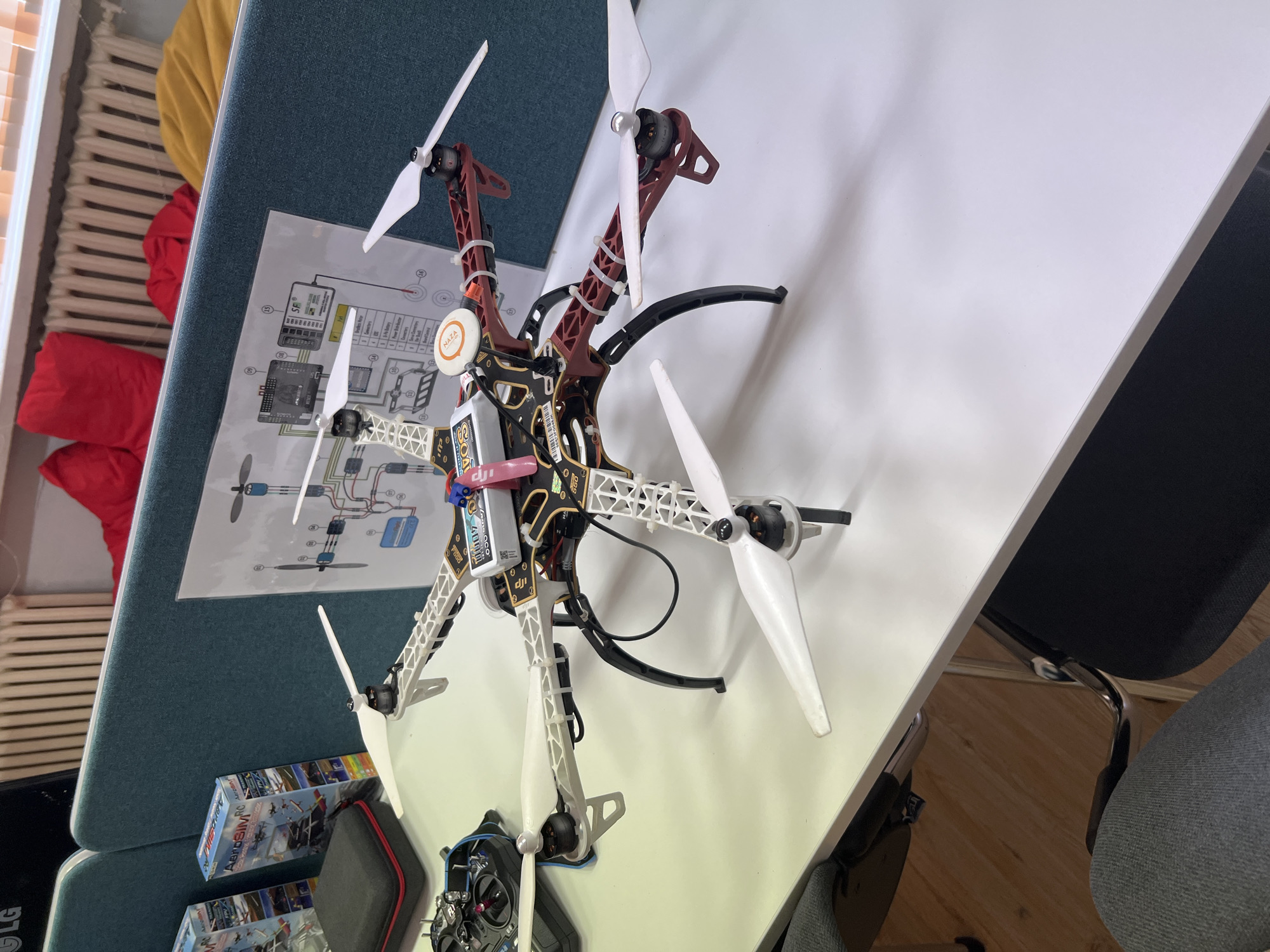 The image depicts a close-up, eye-level view of a drone and its components resting on a white tabletop. The drone, a quadcopter, is centrally positioned and appears to be assembled. It has six white propellers attached to black motor mounts, which are connected to a frame. The frame itself consists of a dark red central body with white structural arms. A white battery pack is secured to the top of the drone's body, with various wires and electronic components visible. A small, orange circular device with "NAZA" written on it is also attached to the drone. To the left of the drone, a black remote controller with a small screen is partially visible. Behind the drone, a blue partition wall is present, adorned with a printed diagram of electronic circuitry, likely related to drone components. Two boxes with graphics of aircraft and the text "AeroSIM RC" are also visible on the left. The background shows a radiator and some colorful cushions on a shelf. The scene is indoors, possibly an office or workshop environment, given the presence of partitions and a desk. There are no people visible in the image. The lighting suggests a well-lit indoor space. The location is specified as the city of Pravets, Bulgaria.