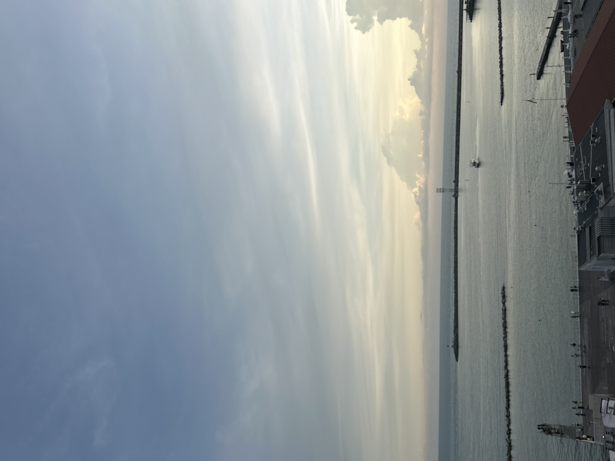 This is an aerial, high-angle, vertical shot of the Black Sea coast in Sochi, Russia, during what appears to be late afternoon or early evening. The sky is partly cloudy with a soft, diffused light. The sea is calm, with a few small boats visible. Two breakwaters extend into the water, one with a lighthouse at its end. In the foreground, there is a promenade with people walking and standing, and some buildings with red roofs. The overall atmosphere is peaceful and serene.