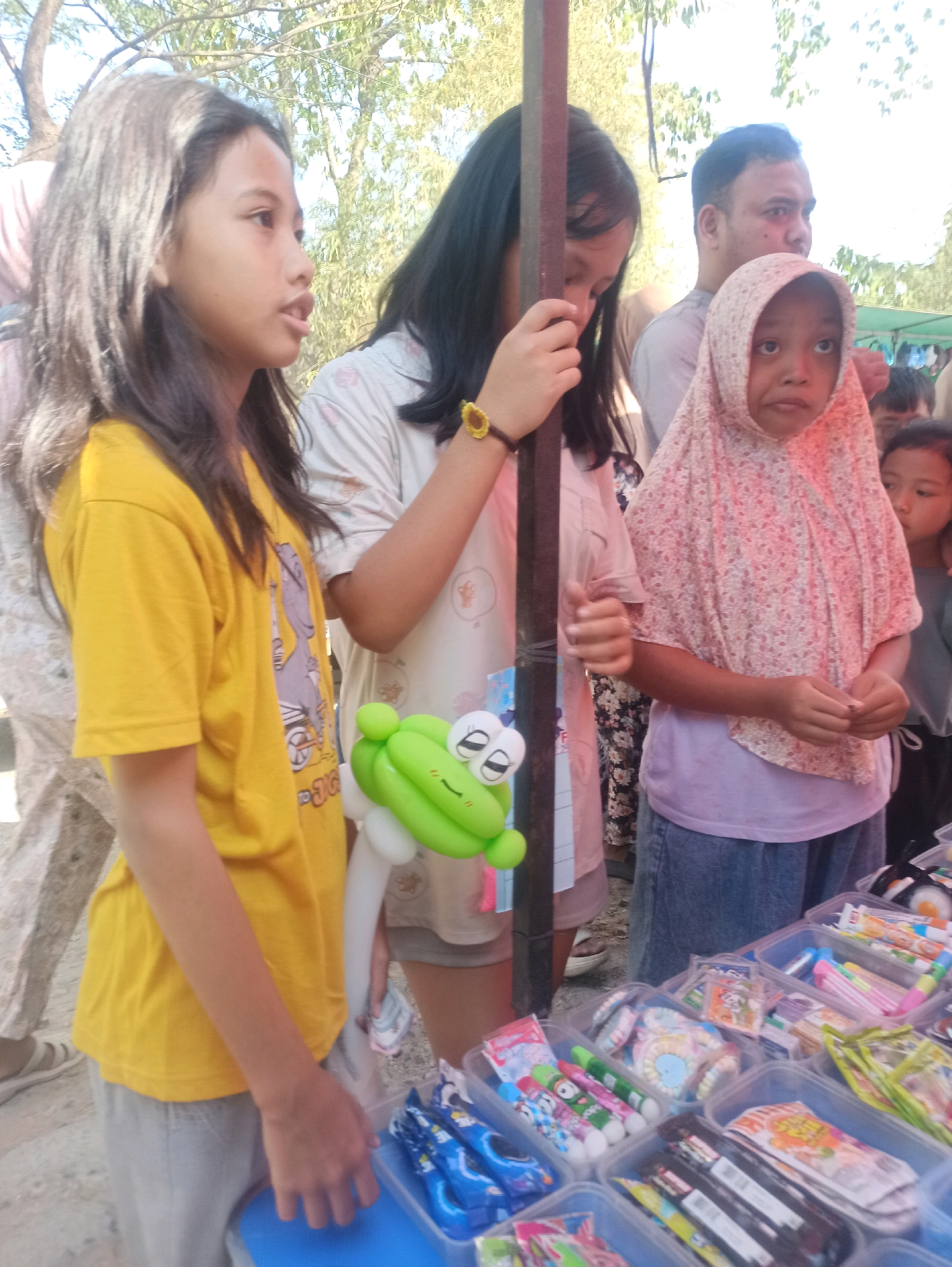 In Telagamurni, Indonesia, at a bustling outdoor market stall, a young girl in a yellow t-shirt stands to the left, her gaze directed away from the camera. Beside her, another girl in a light-colored shirt holds a tall vertical pole, her attention drawn downwards. To the right, a young girl wearing a floral pink hijab and a lavender shirt looks towards the camera with wide eyes. Behind them, a man with short dark hair is partially visible. The foreground displays a variety of colorful items, likely candies and small toys, arranged in clear plastic containers on a table. The overall scene suggests a lively community event or market day.