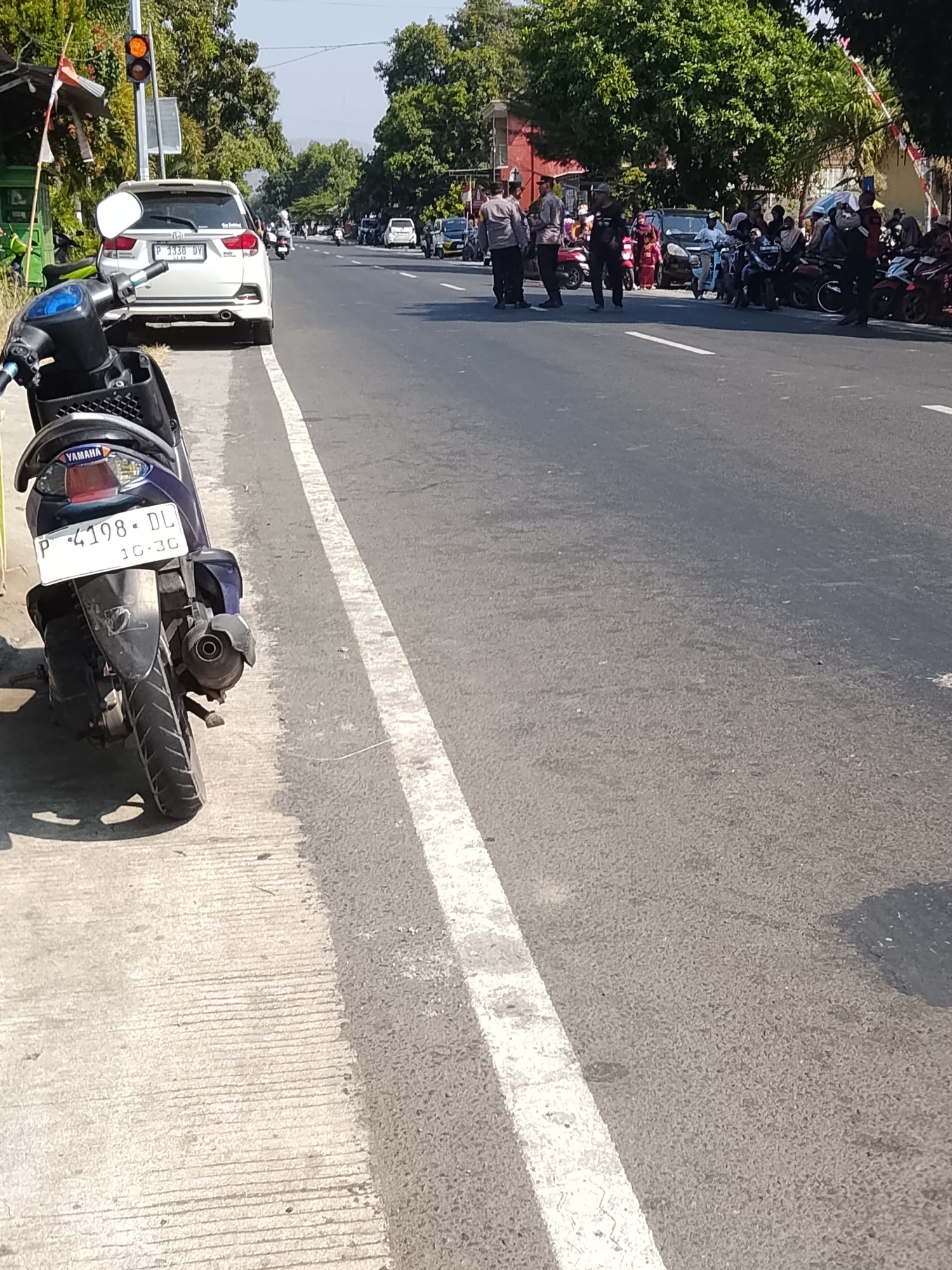 The image depicts a street scene in Buduan, Indonesia, with a focus on a parked purple scooter in the foreground. The scooter has the license plate "P 4198 DL" and "10.30" printed on it. Behind the scooter, on the road, several individuals are visible, some appearing to be police officers in uniform, standing near a group of motorcycles and a white car. Other people, some on motorcycles, are also present on the road. Trees line the side of the road, and there are buildings in the background. The traffic light is visible, showing an orange light. The overall environment suggests a public street in an Indonesian city on a sunny day.