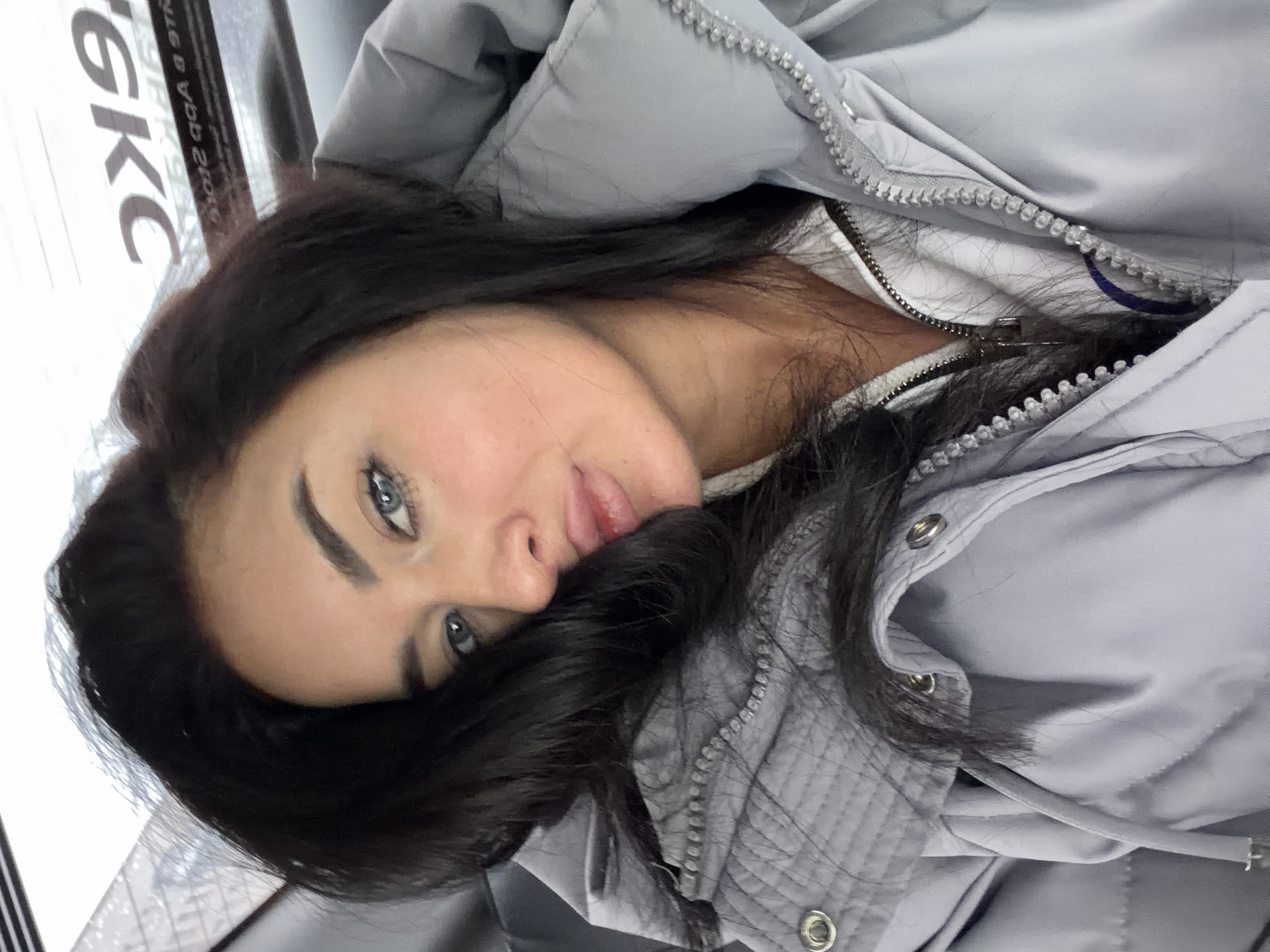 A woman with dark hair and blue eyes lies in what appears to be an indoor setting. She is wearing a light grey quilted jacket. The setting might be a bus, as evidenced by lettering that includes "OKC" visible in the background. No specific location in Moscow, Russia can be determined from the image.