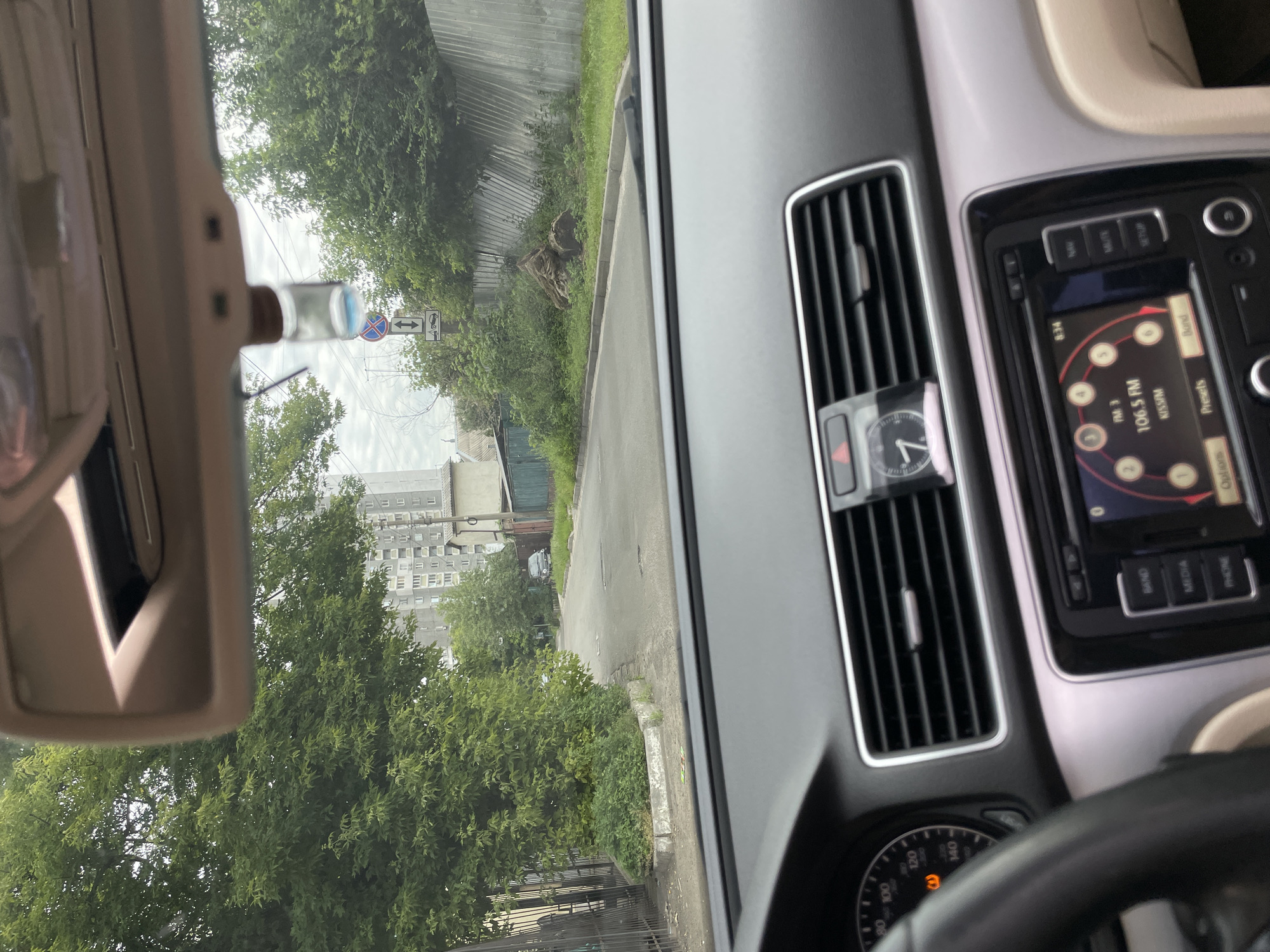 The image is taken from the interior of a car, looking forward through the windshield. The dashboard is visible in the foreground, with a steering wheel on the left and a radio display in the center. Beyond the car, a paved road curves to the left, lined with trees and a fence on the right. Residential buildings and more trees are visible in the background. The sky is overcast. The scene appears to be a street in a residential area of Kyiv, Ukraine, possibly on a cloudy day.